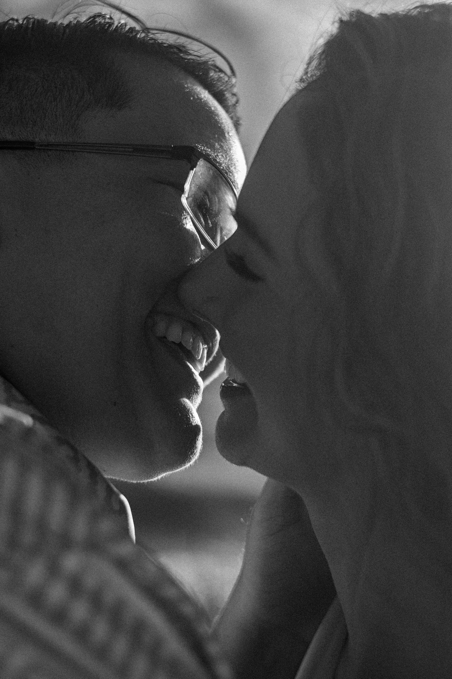 Black and white close-up of a smiling man with glasses and a woman with long hair about to kiss in soft, warm backlight.