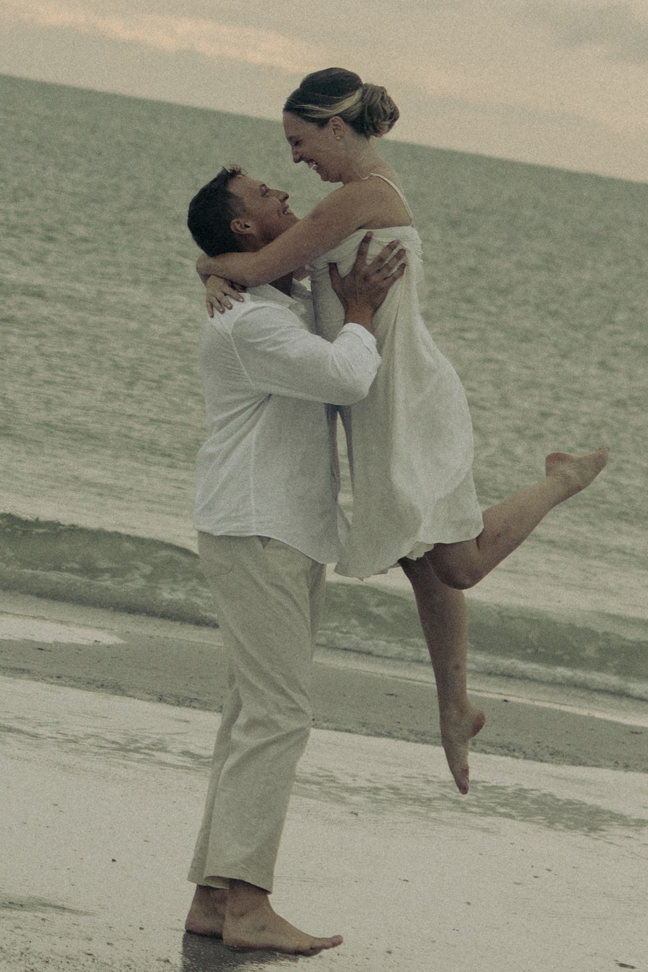 Barefoot man in white lifts a smiling woman in a white dress into the air on a quiet beach at sunset.