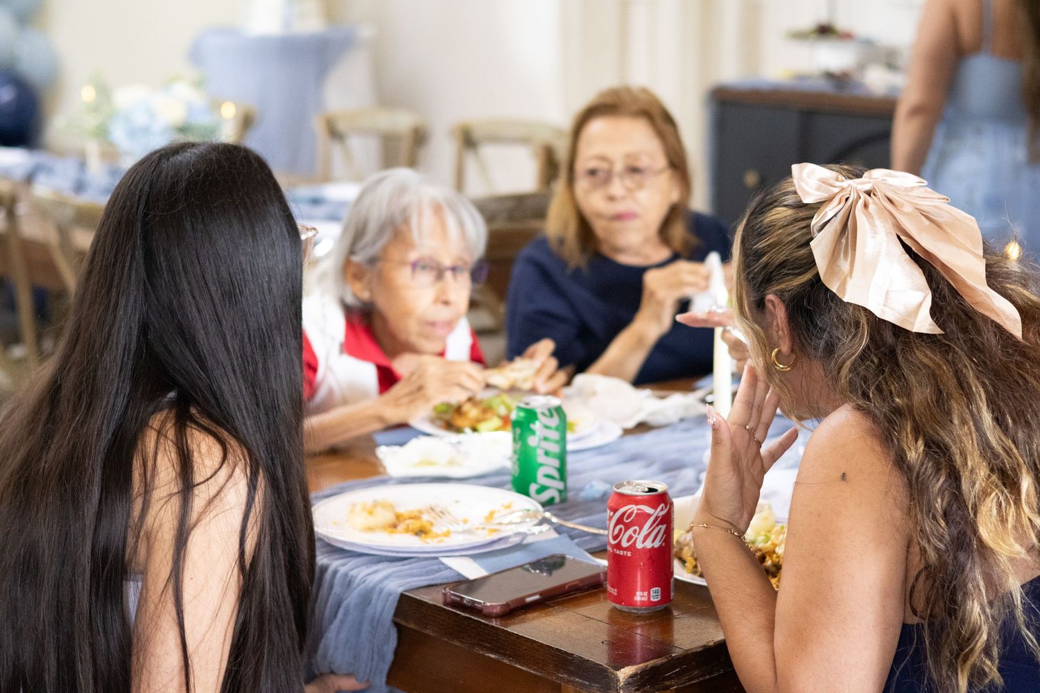 Guests share a meal at a baby shower table with plates of food and soda cans, including an older woman smiling.
