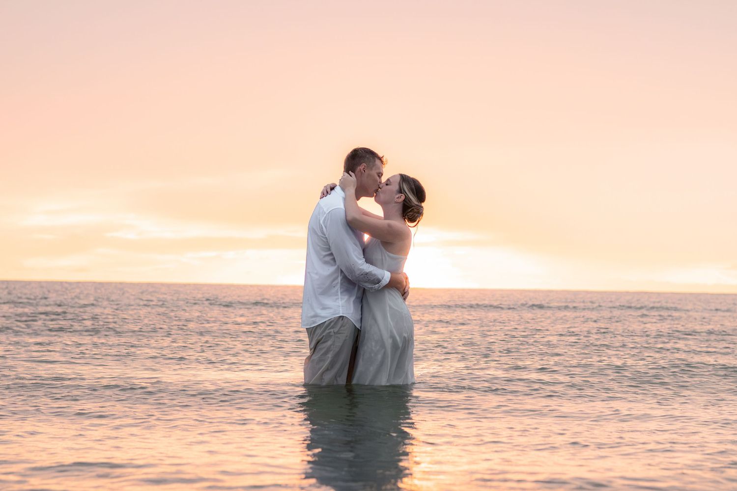 Couple shares a kiss while standing waist-deep in calm ocean water beneath a glowing pink and orange sunset sky.