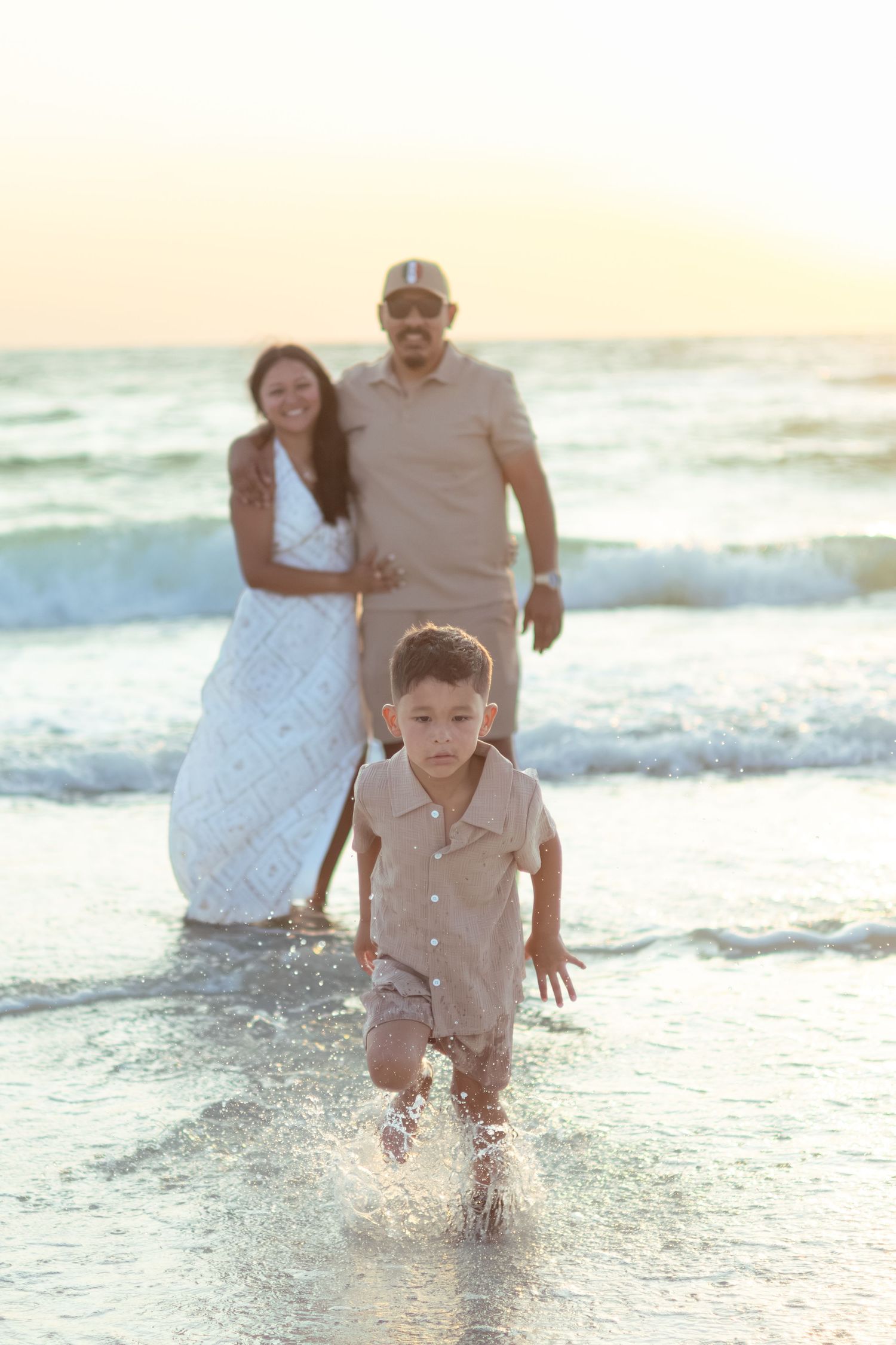 Smiling couple embraces in the ocean surf at sunset while their young son runs and splashes through the shallow water toward the camera.