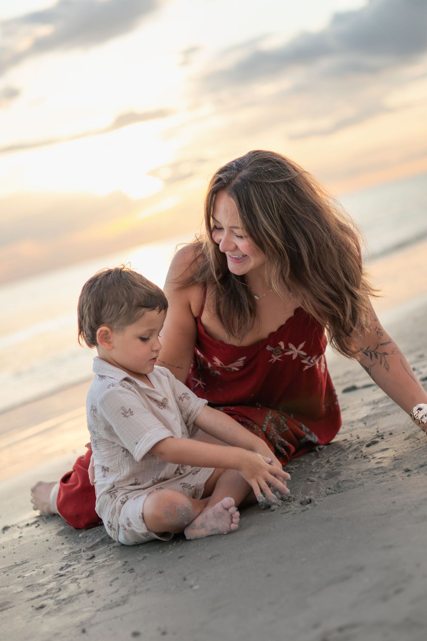 A smiling mother in a red dress sits on the sand beside her young son at sunset, playing together by the ocean.