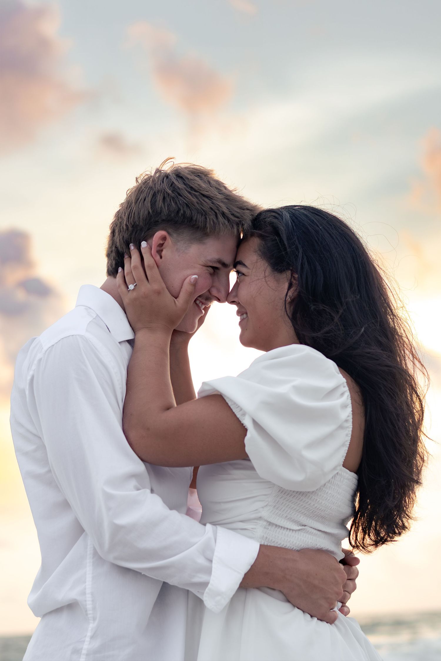An engaged couple in white touches foreheads and smiles in a tender embrace on the beach, her diamond ring visible.
