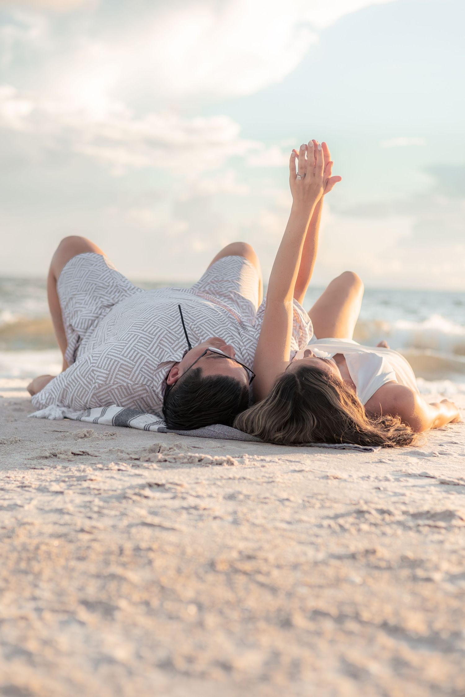 Couple lying on their backs on a sandy beach at sunset, reaching their hands toward the cloudy sky with ocean waves behind them.