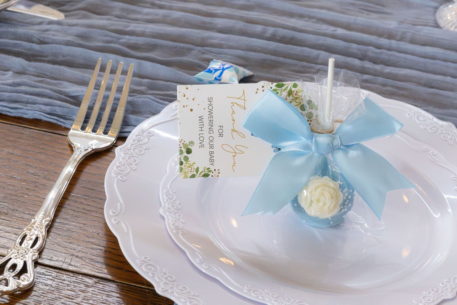 Place setting with a white plate, blue bow favor, thank-you card and silver fork on a gray table runner at a baby shower.