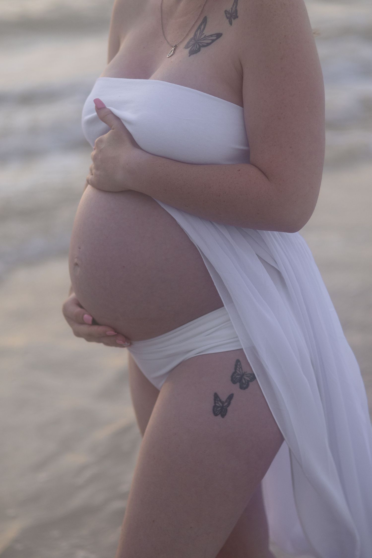 Pregnant woman in white fabric holds her belly on the shore, showing butterfly tattoos on her chest and thigh.