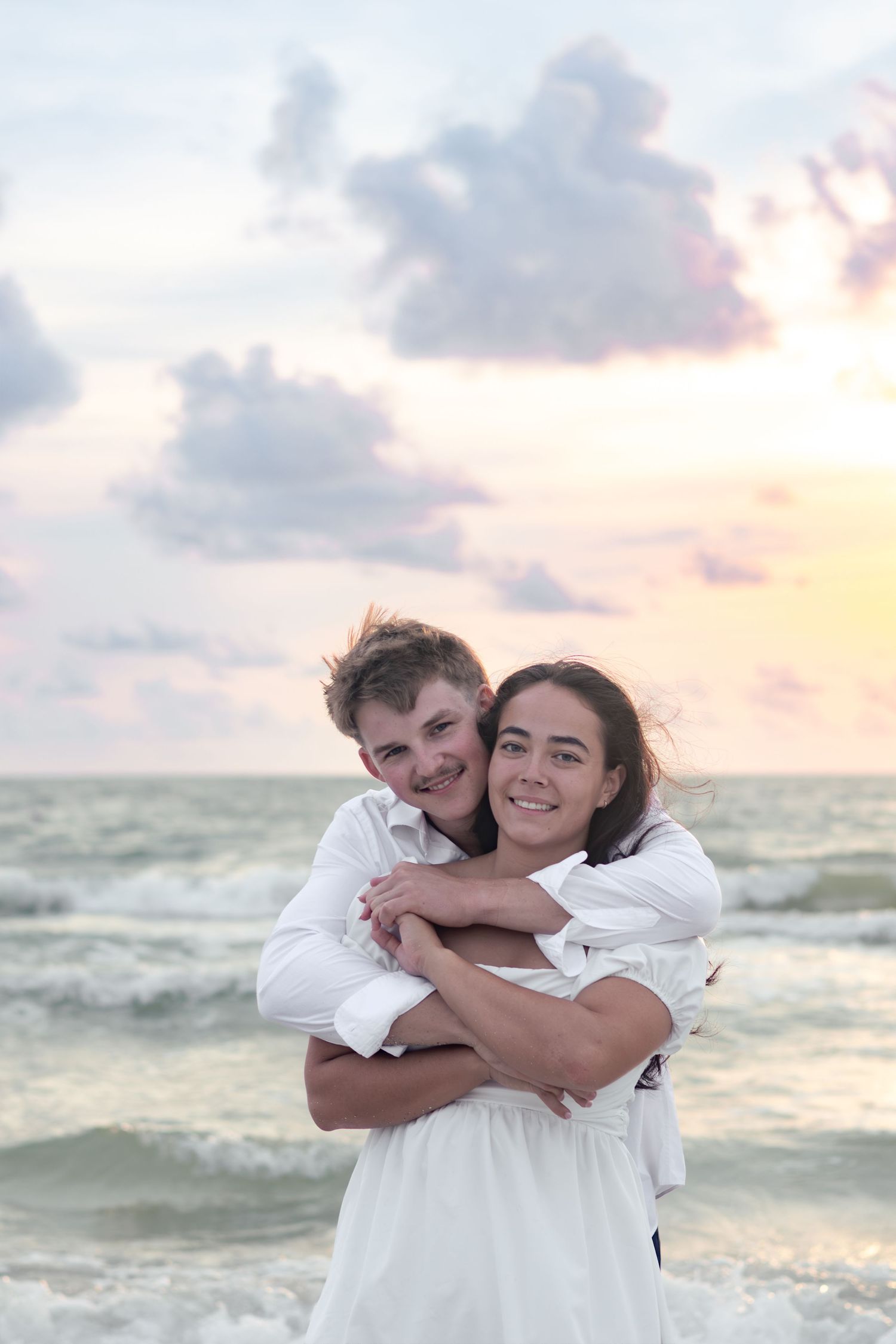 Smiling man hugging a woman from behind on the beach with pastel sunset clouds over the ocean.