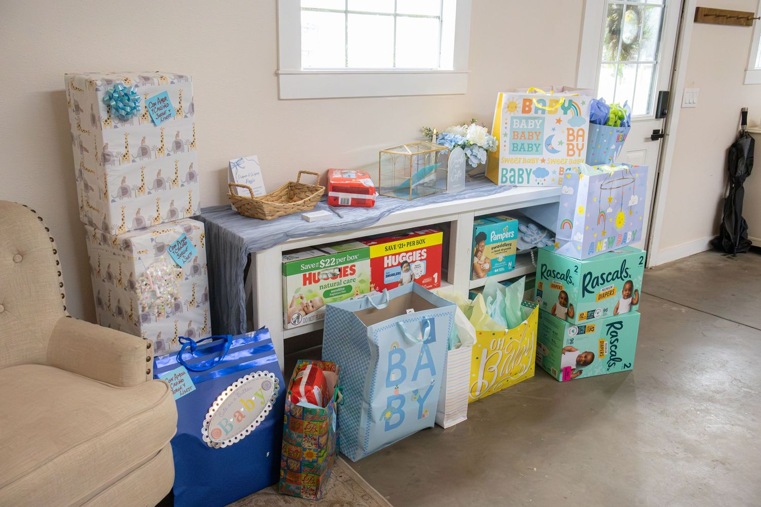 Stacks of wrapped baby shower gifts, bags and presents fill a table and floor beside a cozy armchair.