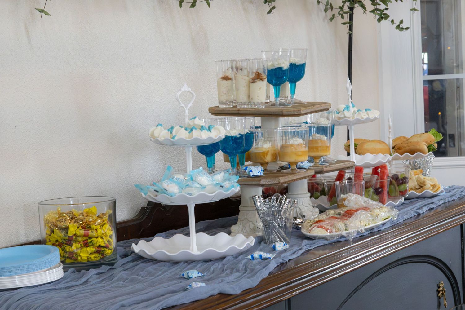 Baby shower dessert table with tiered stands, blue glass cups, treats and snacks on a gray runner over a wooden sideboard.