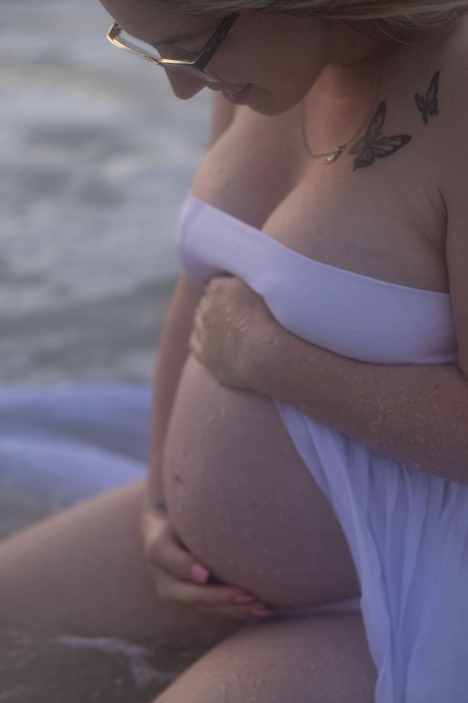 Pregnant woman in glasses and a white wrap cradles her belly while seated at the water's edge during a beach maternity photo session.