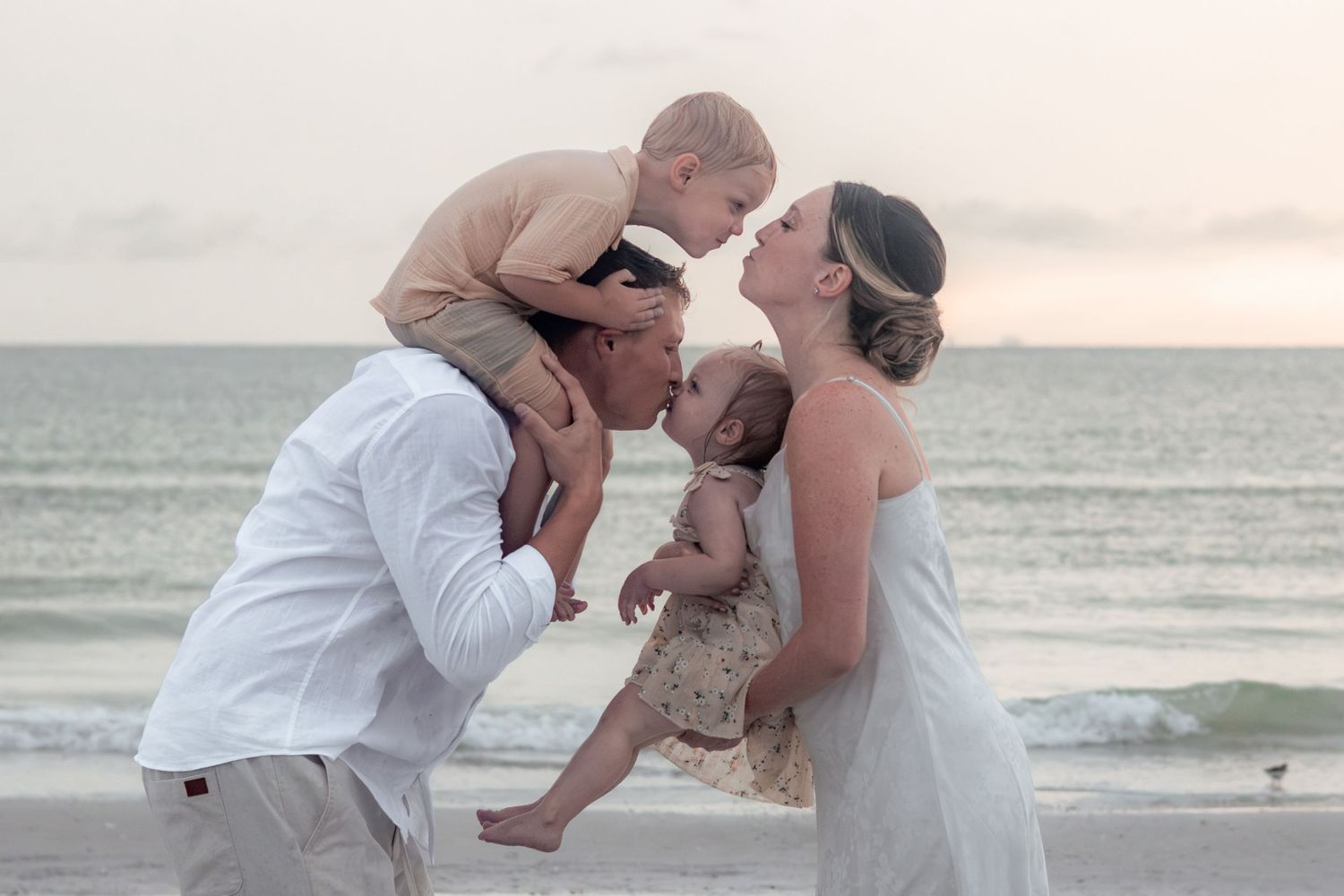 Parents share a kiss on the beach while holding their young son and baby daughter between them near the ocean.
