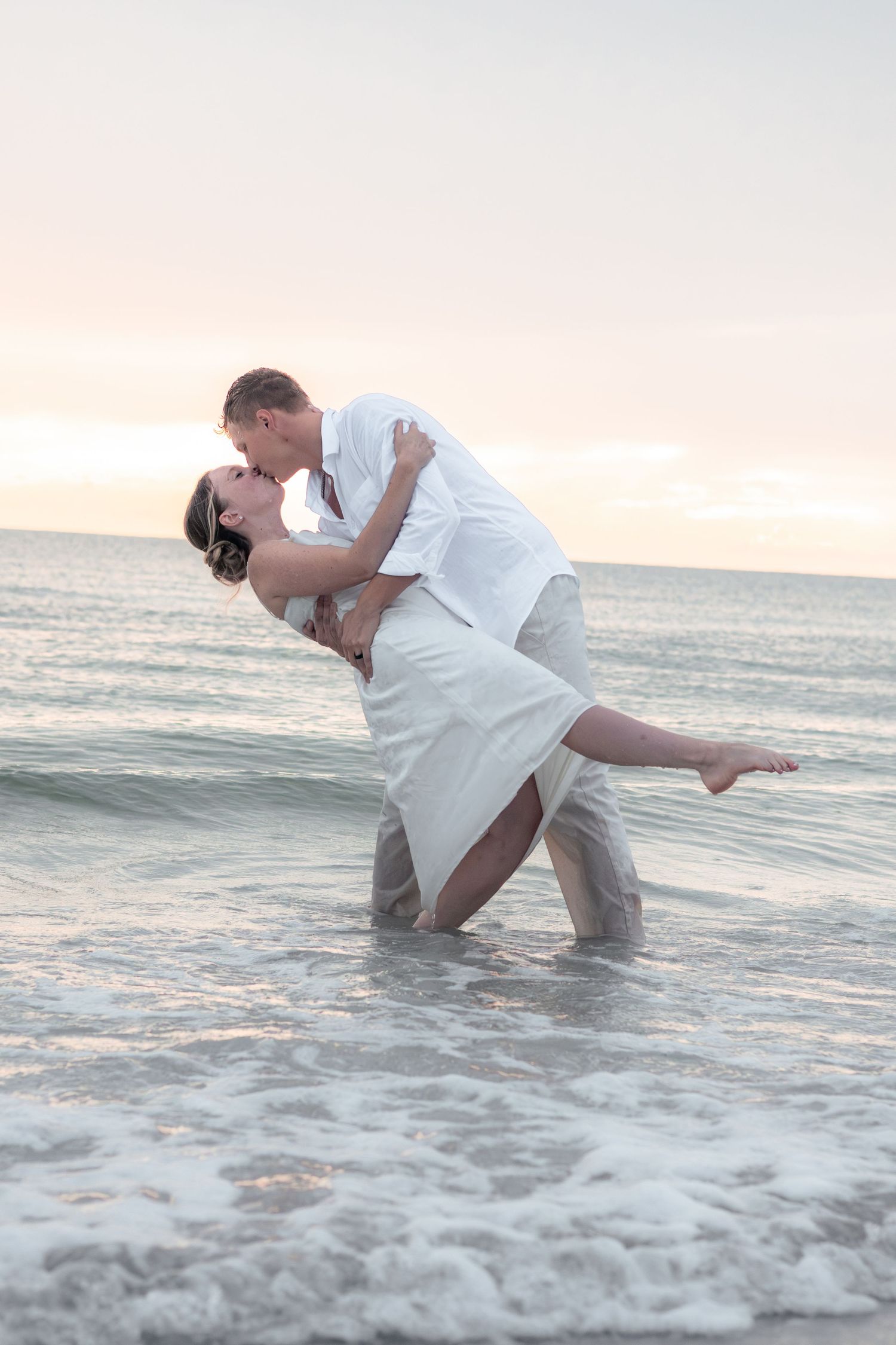 A couple in white shares a romantic dipped kiss while standing in shallow ocean waves at sunset on the beach.