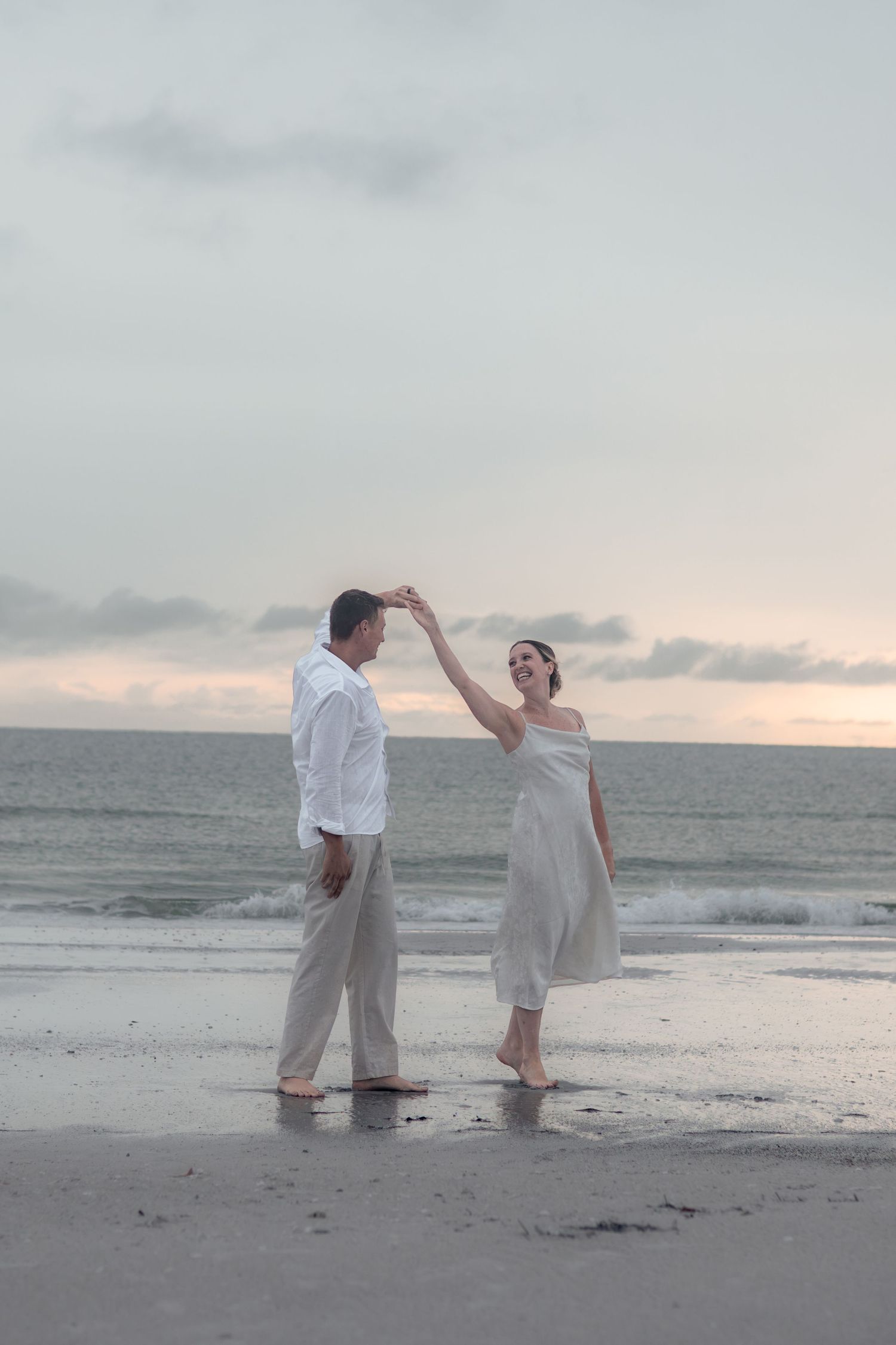 Barefoot man twirls his smiling partner in a white dress as they dance together on the beach near gentle ocean waves.