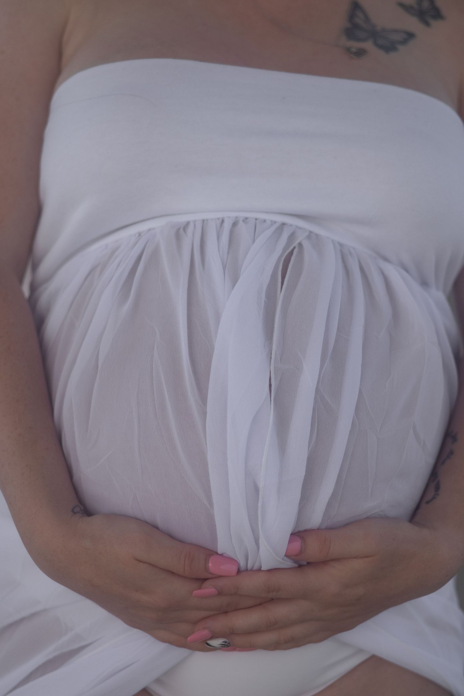 Close-up of a pregnant belly wrapped in sheer white fabric with hands cradled below and a butterfly tattoo on the chest.