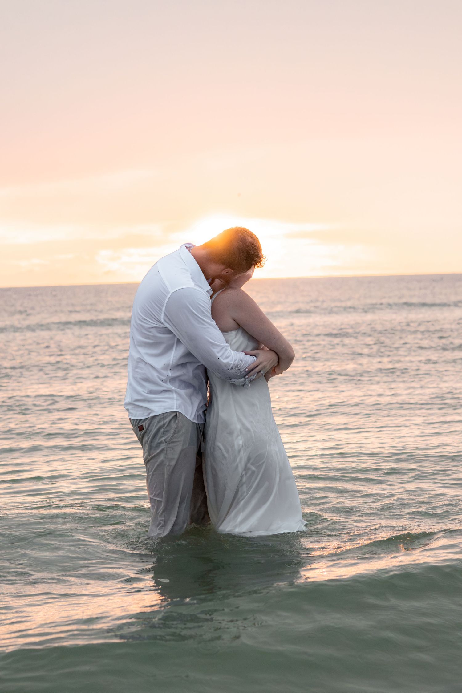 Couple embracing in knee-deep ocean water at sunset, the man in a white shirt and the woman in a white dress, with a pastel sky behind.