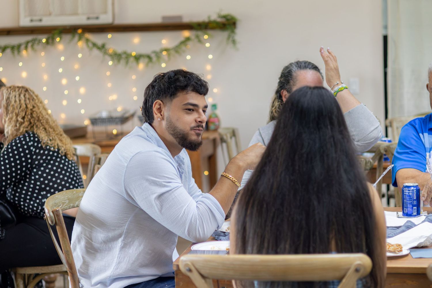 Smiling bearded man in a light blue shirt chats with guests seated at a wooden table under string lights.