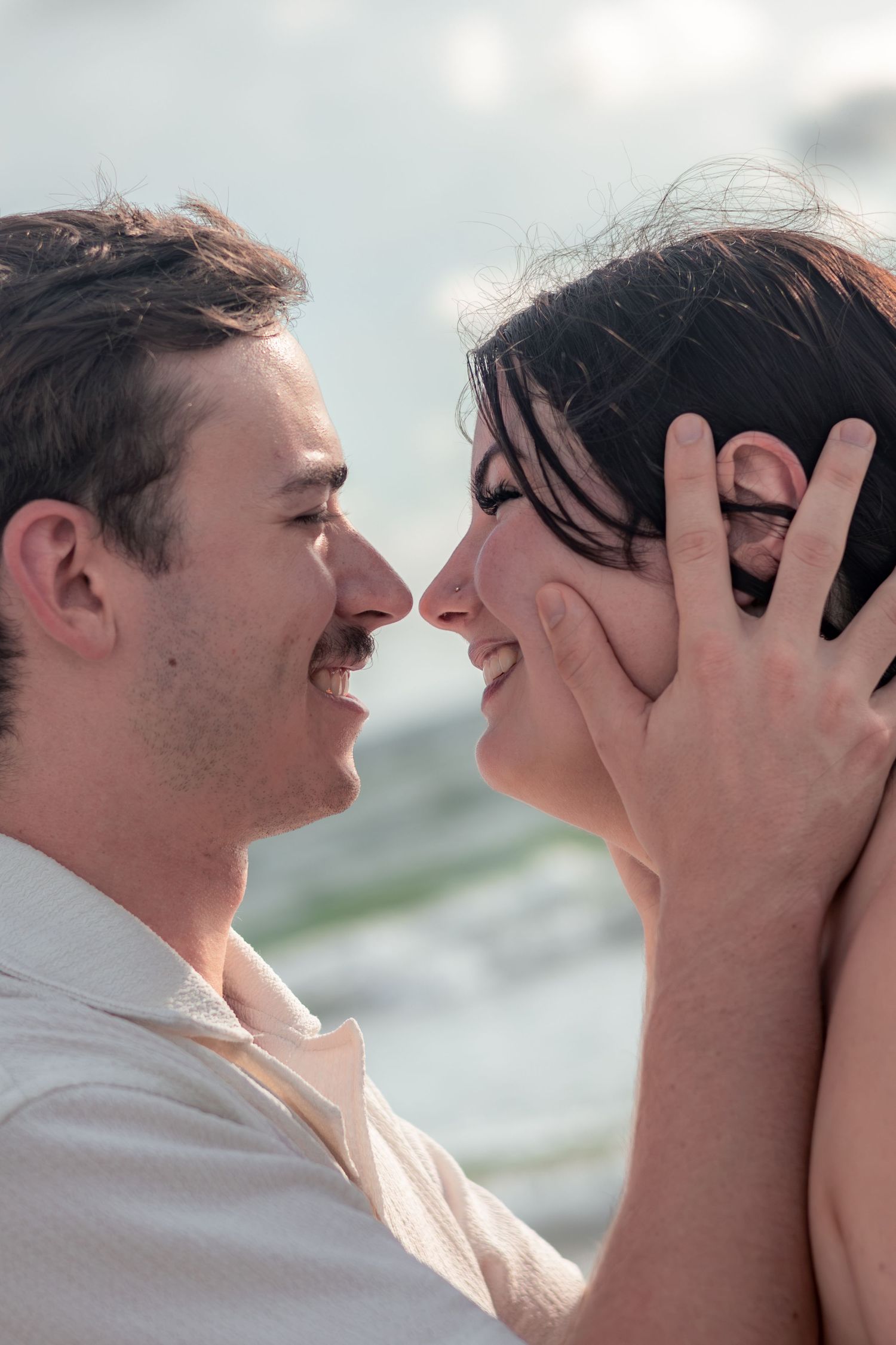 Smiling couple touching noses in an intimate moment on the beach, her hand gently cradling his face with ocean waves behind them.