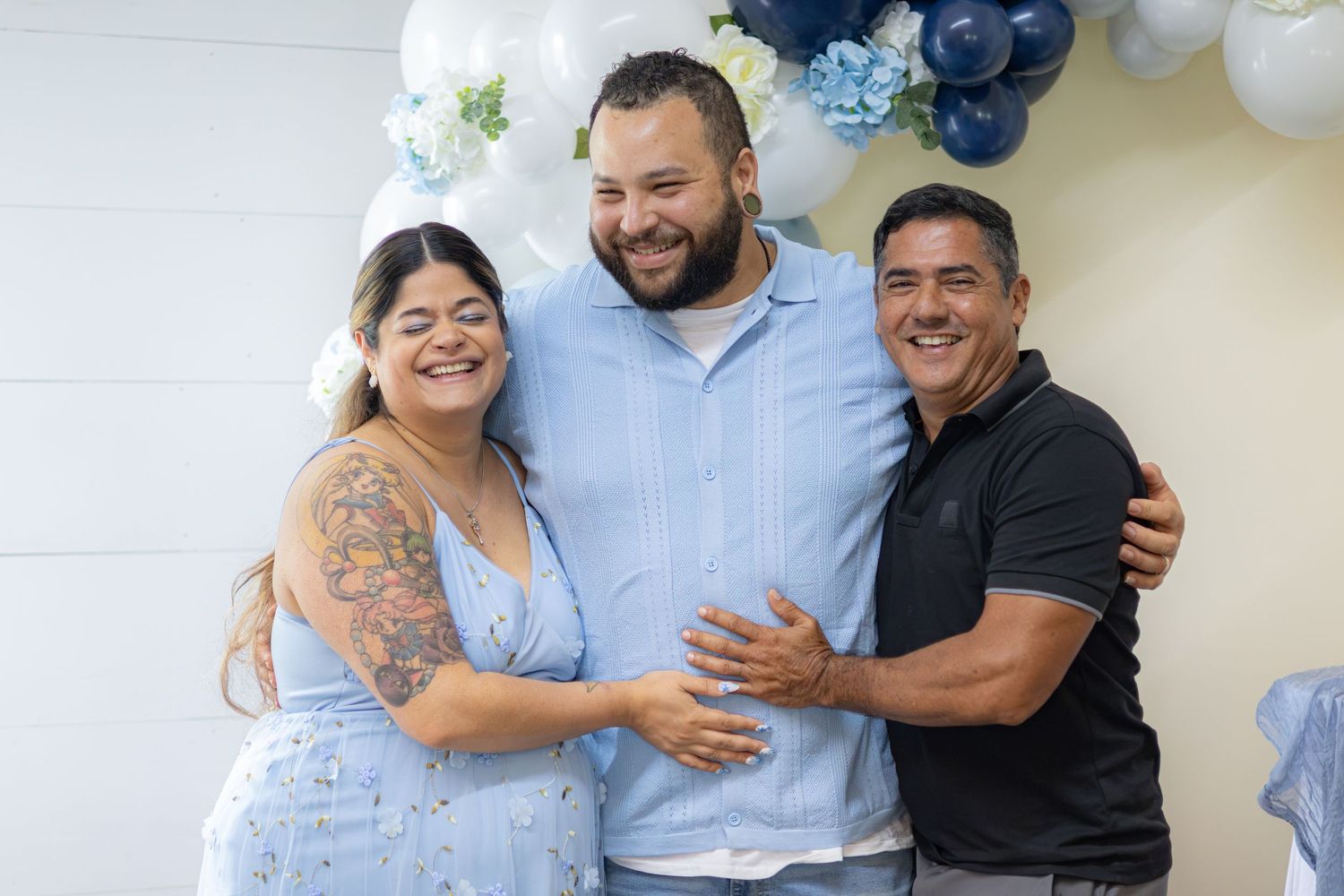Smiling woman in a blue dress and two men in blue and black shirts embrace in front of balloons at a baby shower.