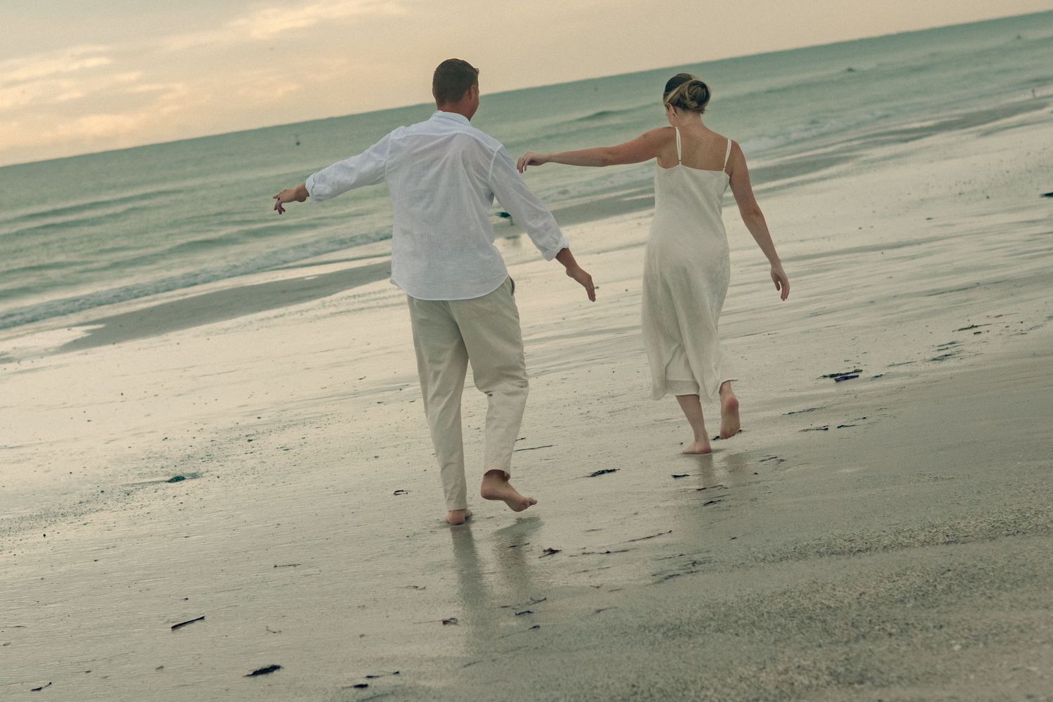 A couple in white beachwear walks barefoot with arms outstretched along the wet sand beside the ocean at dusk.