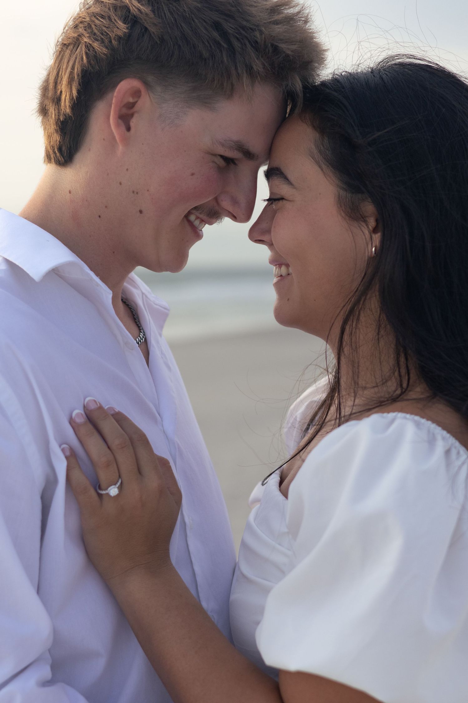 A close-up of a happy couple touching foreheads and smiling on the beach, with her sparkling engagement ring on his chest.