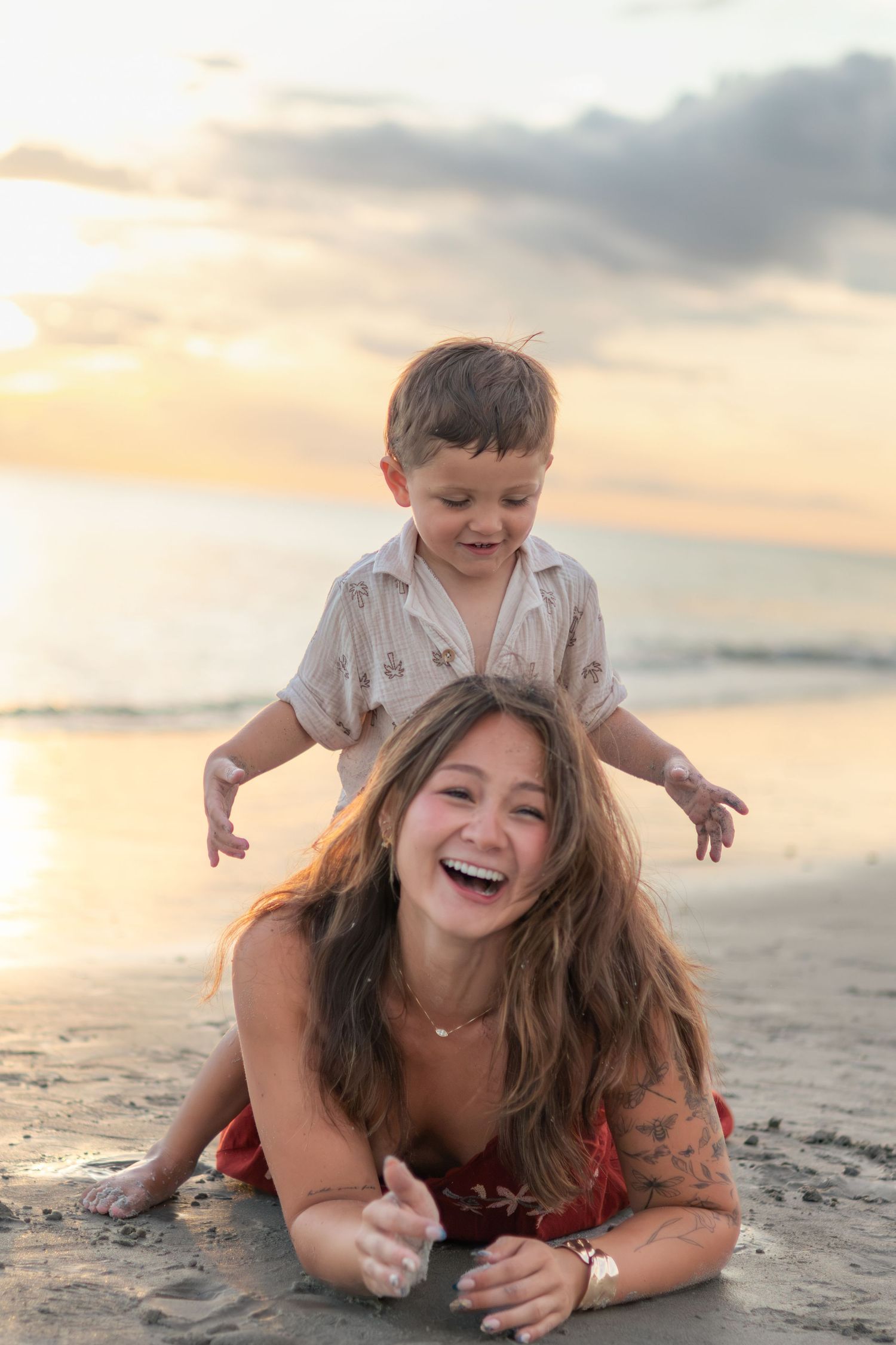 A laughing woman lies on the sandy beach while a happy toddler boy plays on her back at sunset by the ocean.