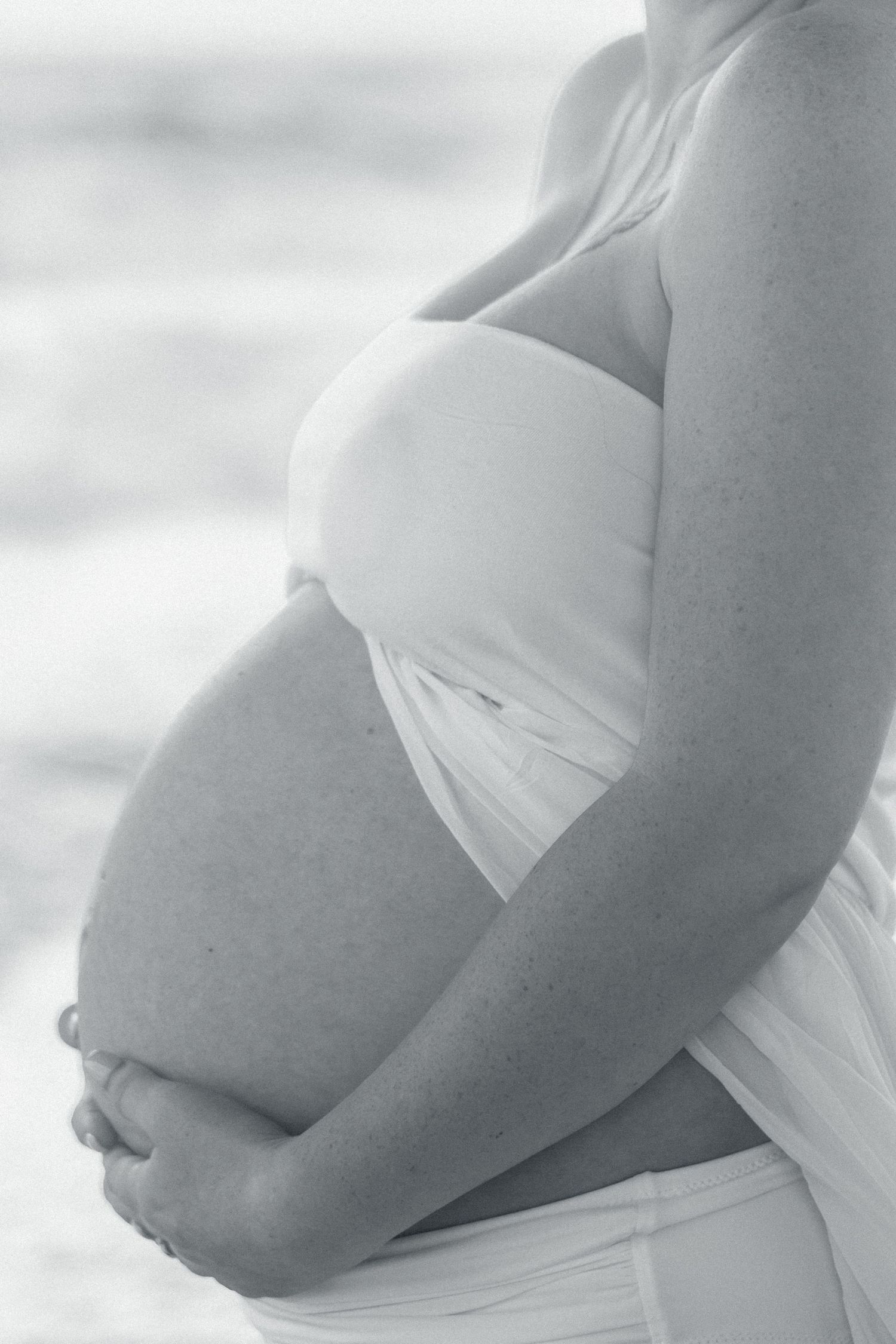Black and white side profile of a pregnant woman in white fabric gently cradling her baby bump near the beach.