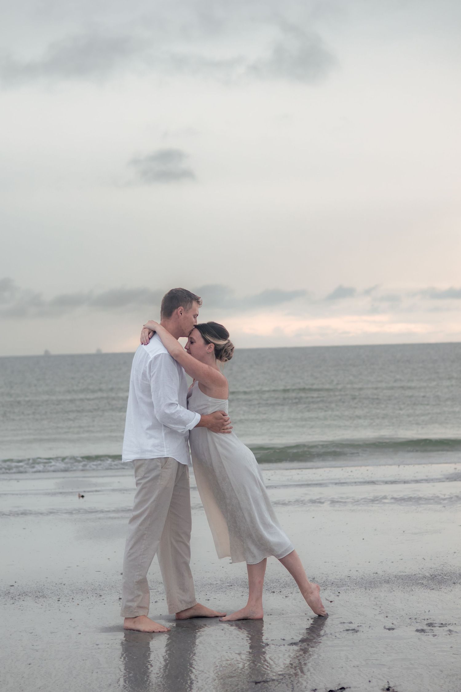 Barefoot couple in white and khaki share a tender embrace on a wet sandy beach beneath a soft cloudy evening sky.