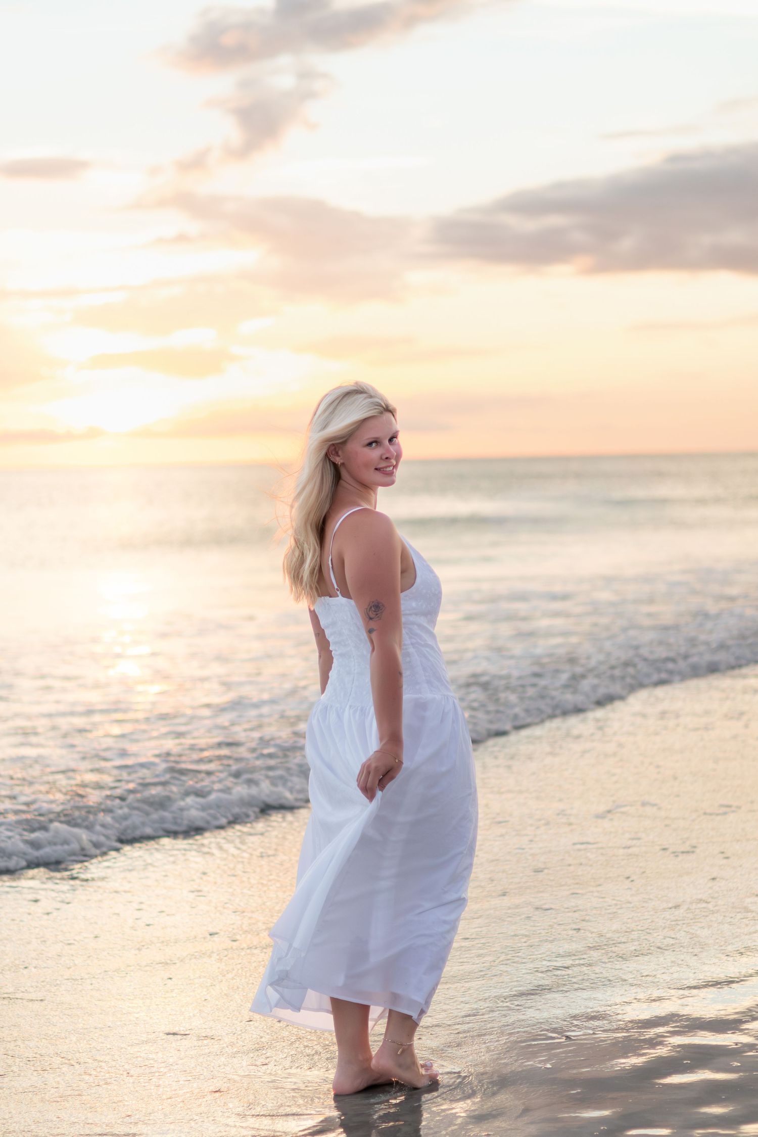 Smiling woman in a flowing white dress stands barefoot on the shore, glancing back as the sun sets over the ocean.