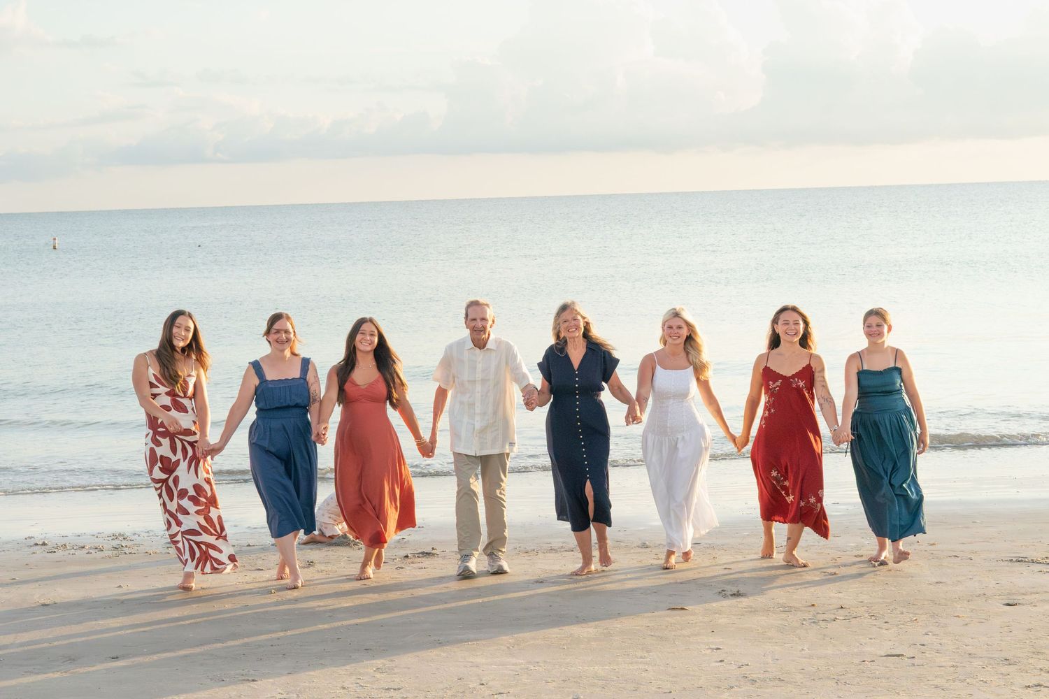 Multigenerational family of women in summer dresses and one man walk hand in hand along the shore at sunset.