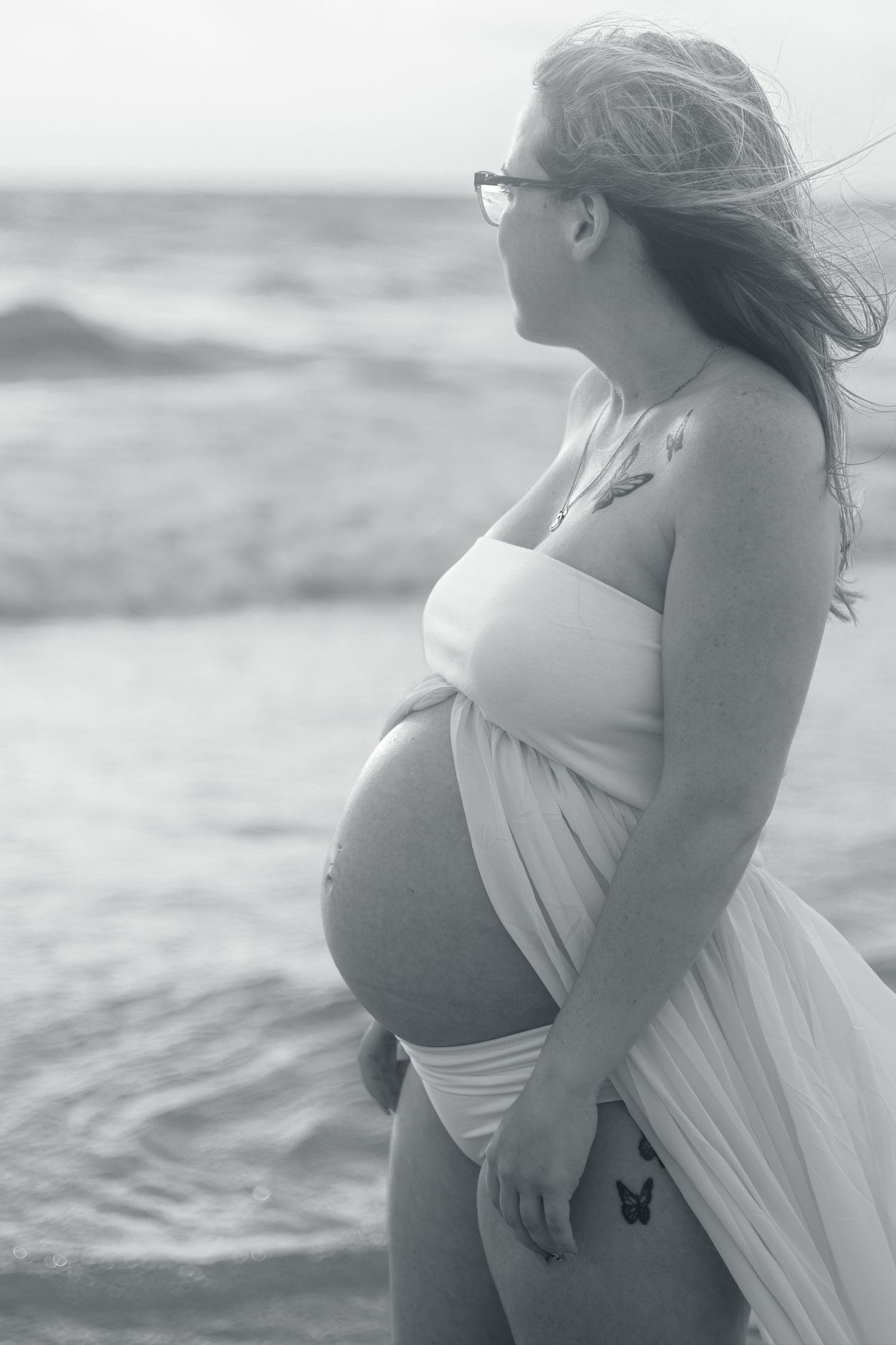 Black and white maternity photo of a pregnant woman in glasses and flowing white fabric gazing out at the ocean waves.