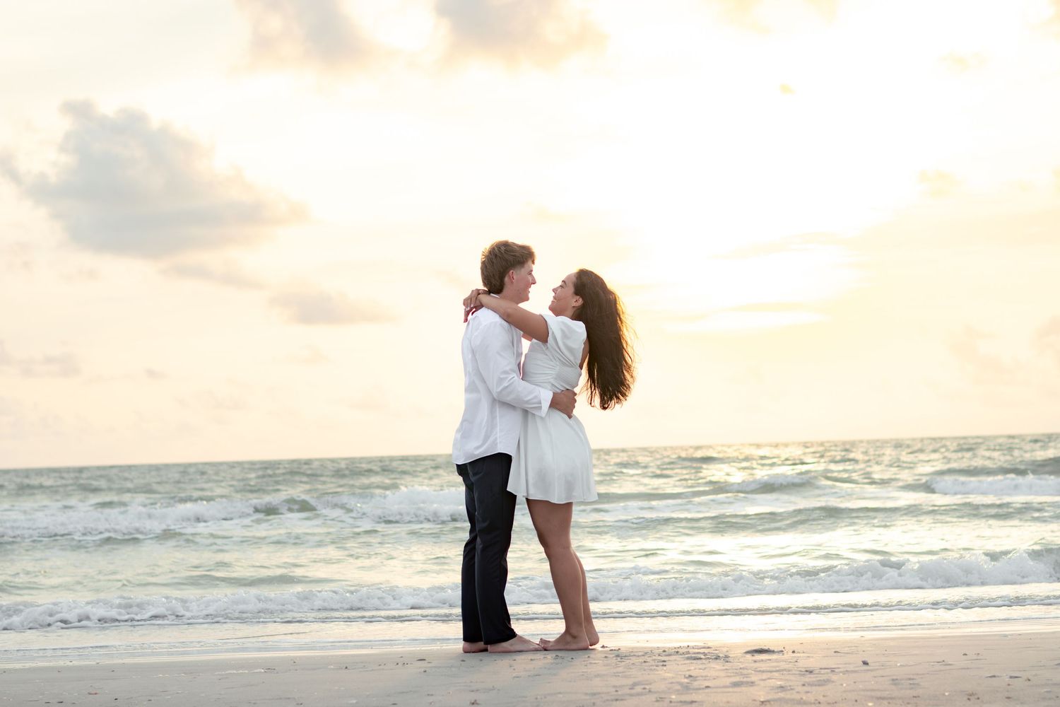 Couple in white embracing and kissing on a sandy beach at sunset with soft waves in the background.