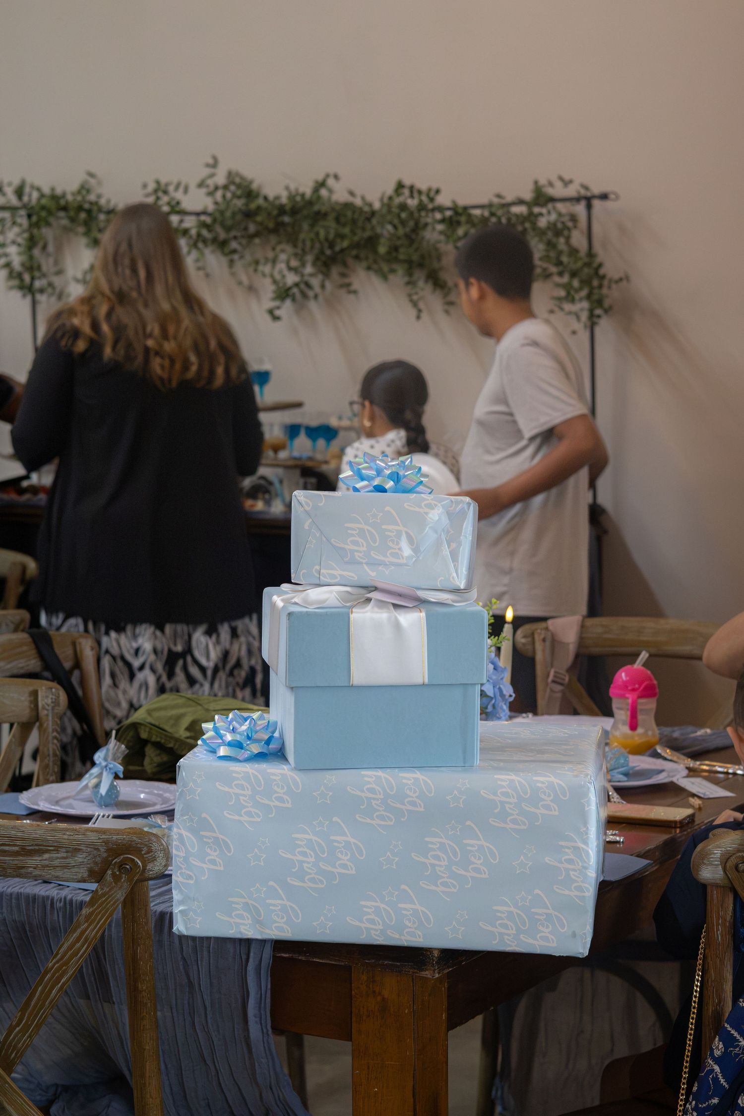 Three stacked light blue gift boxes with bows sit on a rustic table while guests mingle at a baby shower.