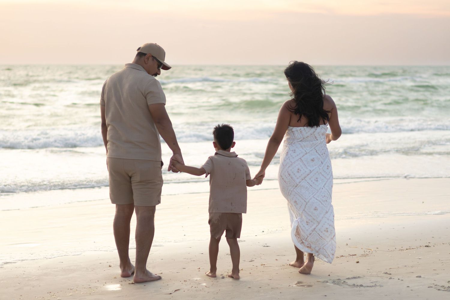 A father, mother and young son hold hands and walk barefoot along the shore toward the ocean at sunset.