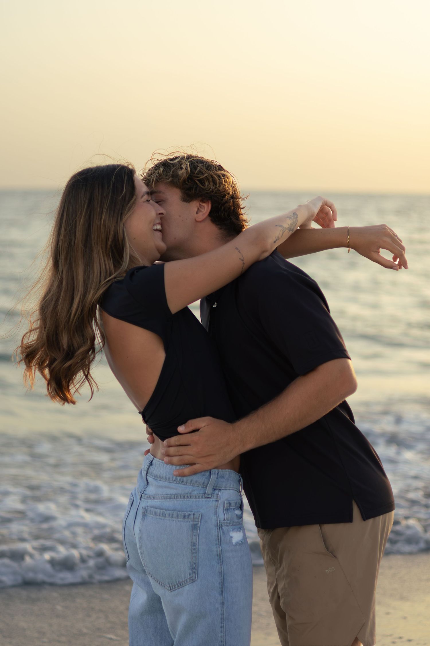 Couple embraces and kisses on the beach at sunset, wearing dark tops and jeans beside gentle waves.