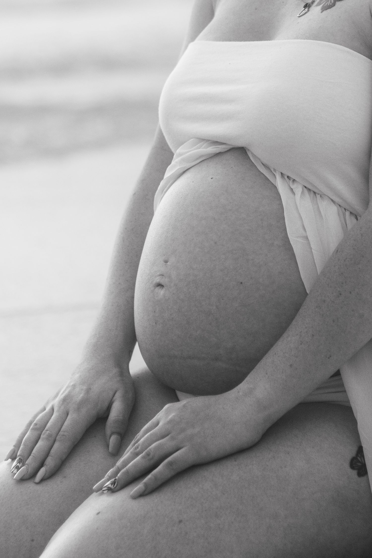 Black and white maternity photo of a seated pregnant woman with both hands resting gently on her bare belly.