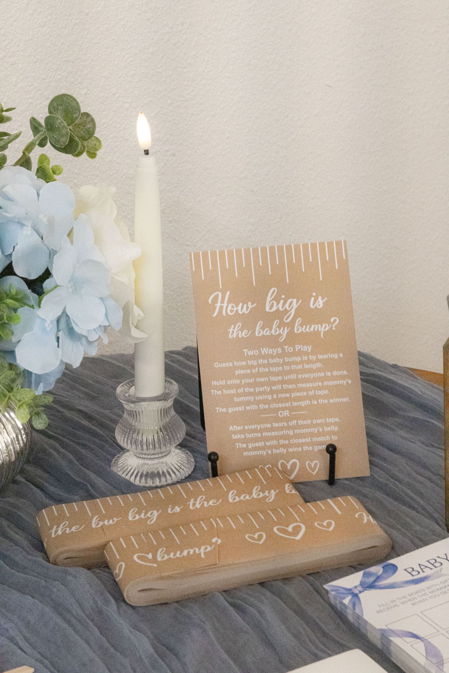 Baby shower game sign and wooden ruler strips for guessing baby bump size beside a lit candle and blue floral arrangement.
