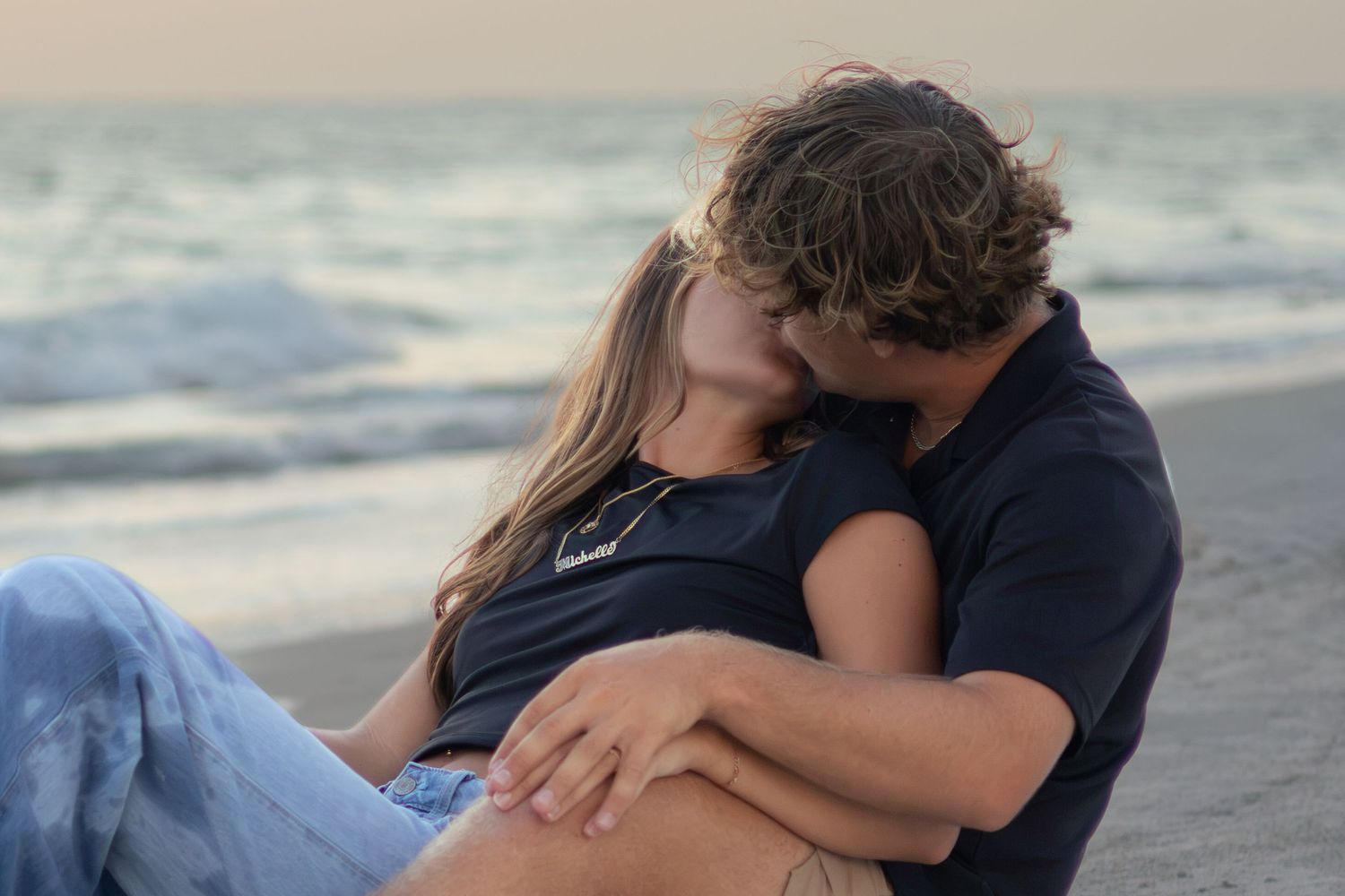 Couple cuddles and kisses on the sand while ocean waves roll in behind them at dusk.
