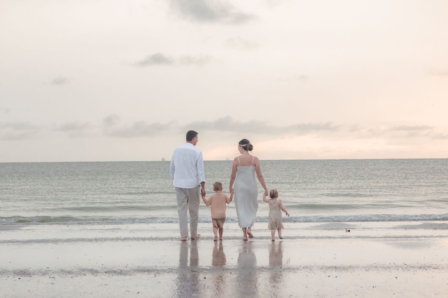 Family of four holds hands and walks toward the ocean at sunset, seen from behind on a wide, calm beach.