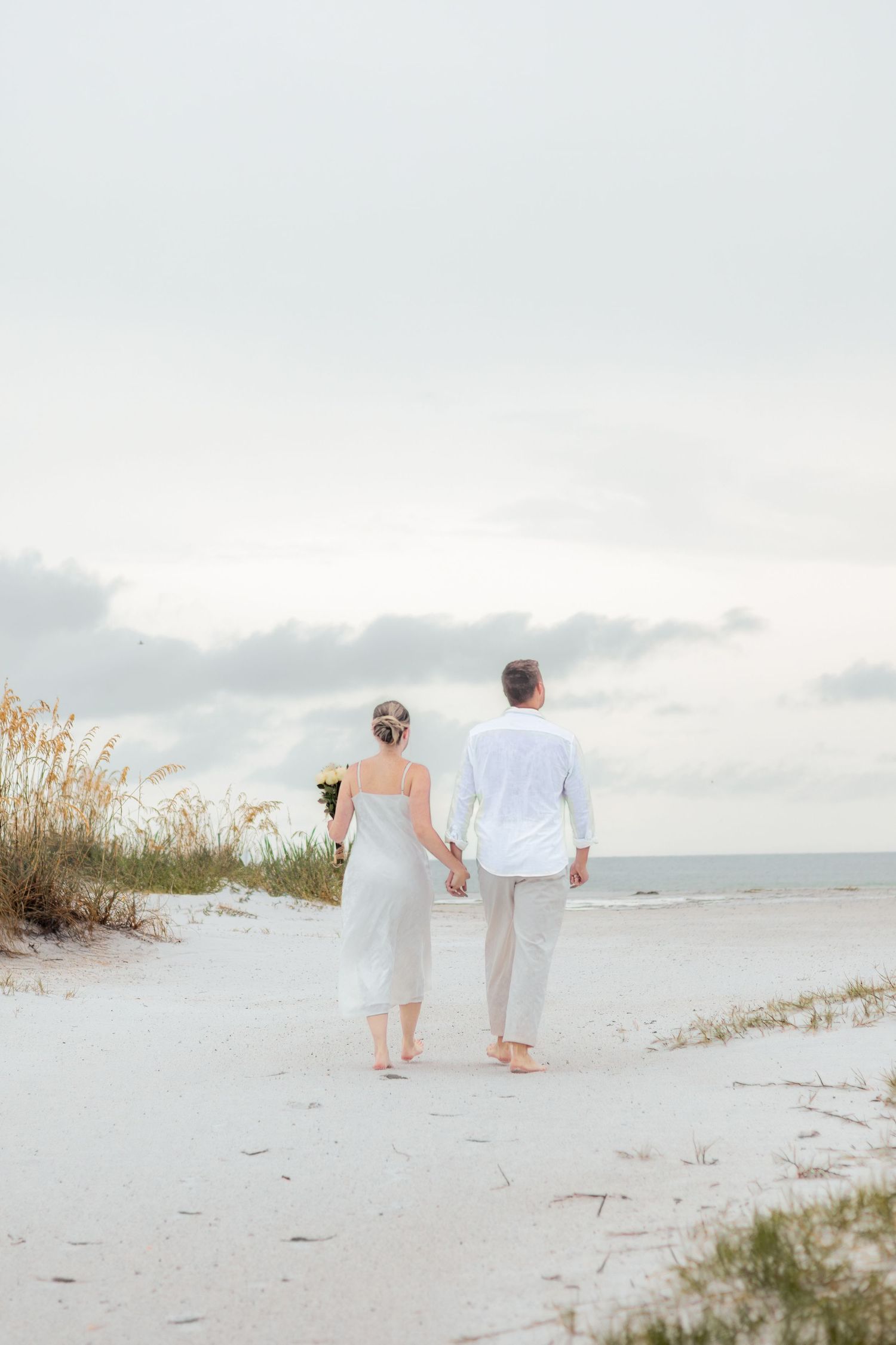 Couple in white outfits holds hands and walks barefoot on a white sand beach toward the ocean.