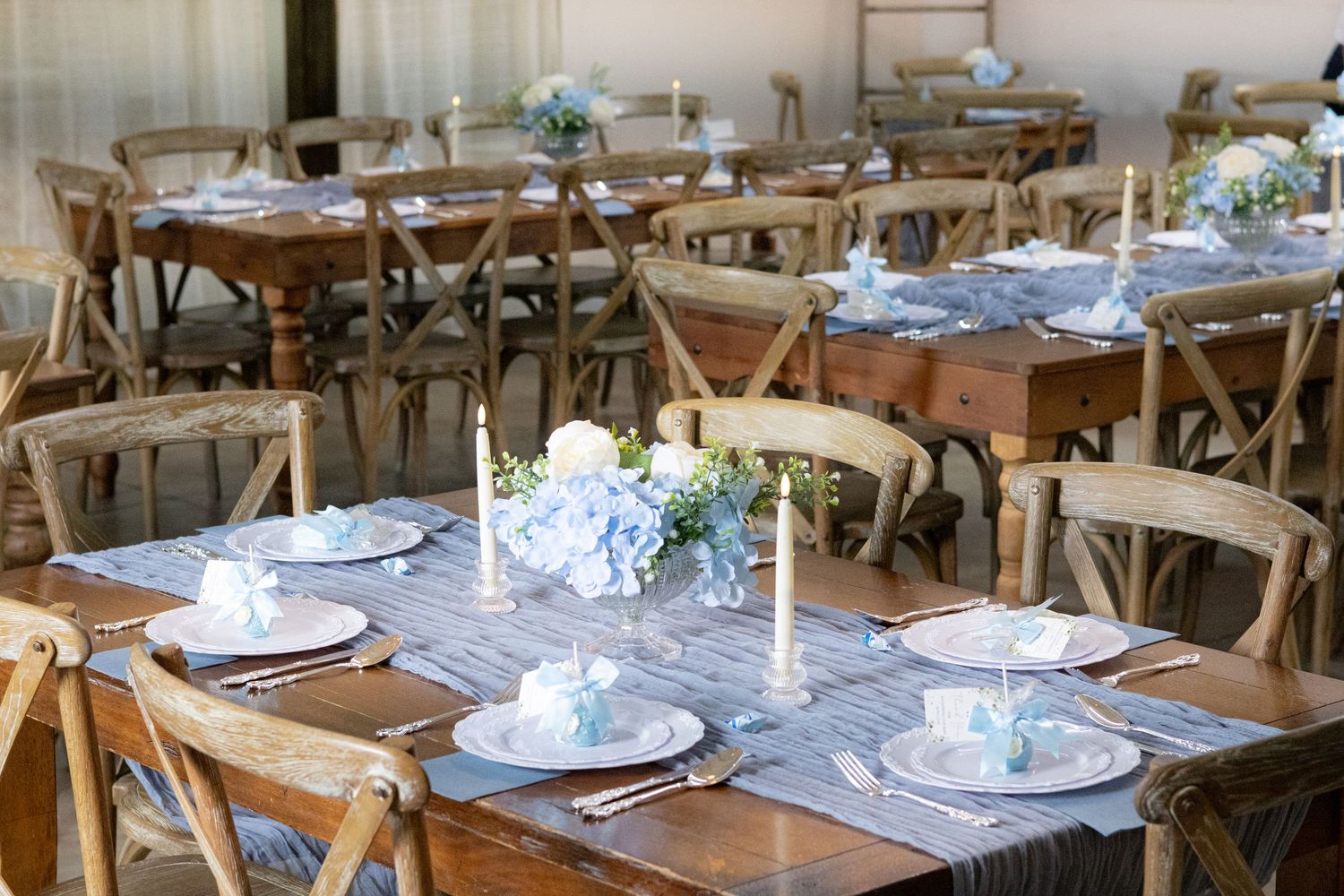 Long rustic wooden tables with gray runners, blue hydrangea centerpieces and candles set for a baby shower reception.