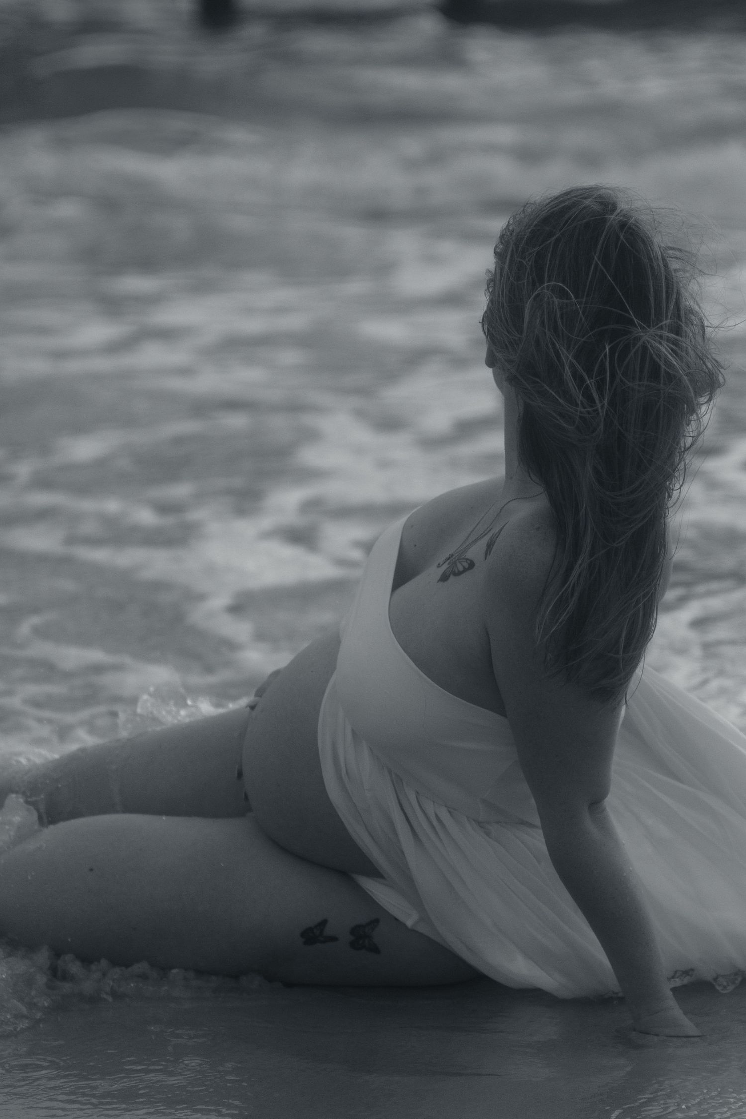 Black and white maternity portrait of a woman sitting in the ocean surf, looking out to sea with her hair in a ponytail.