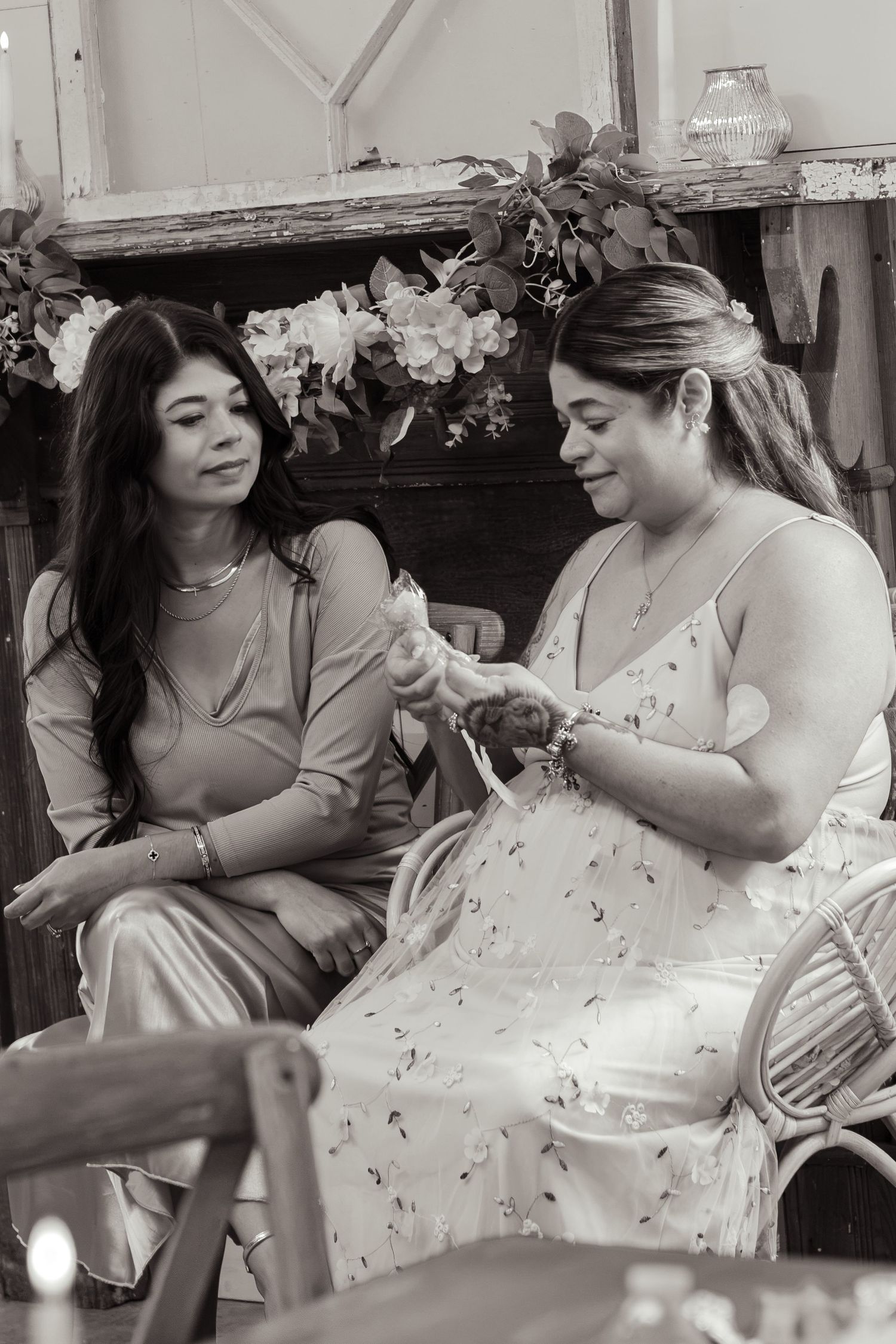 Black and white photo of two women seated together at a baby shower, one holding a small baby item in her hands.