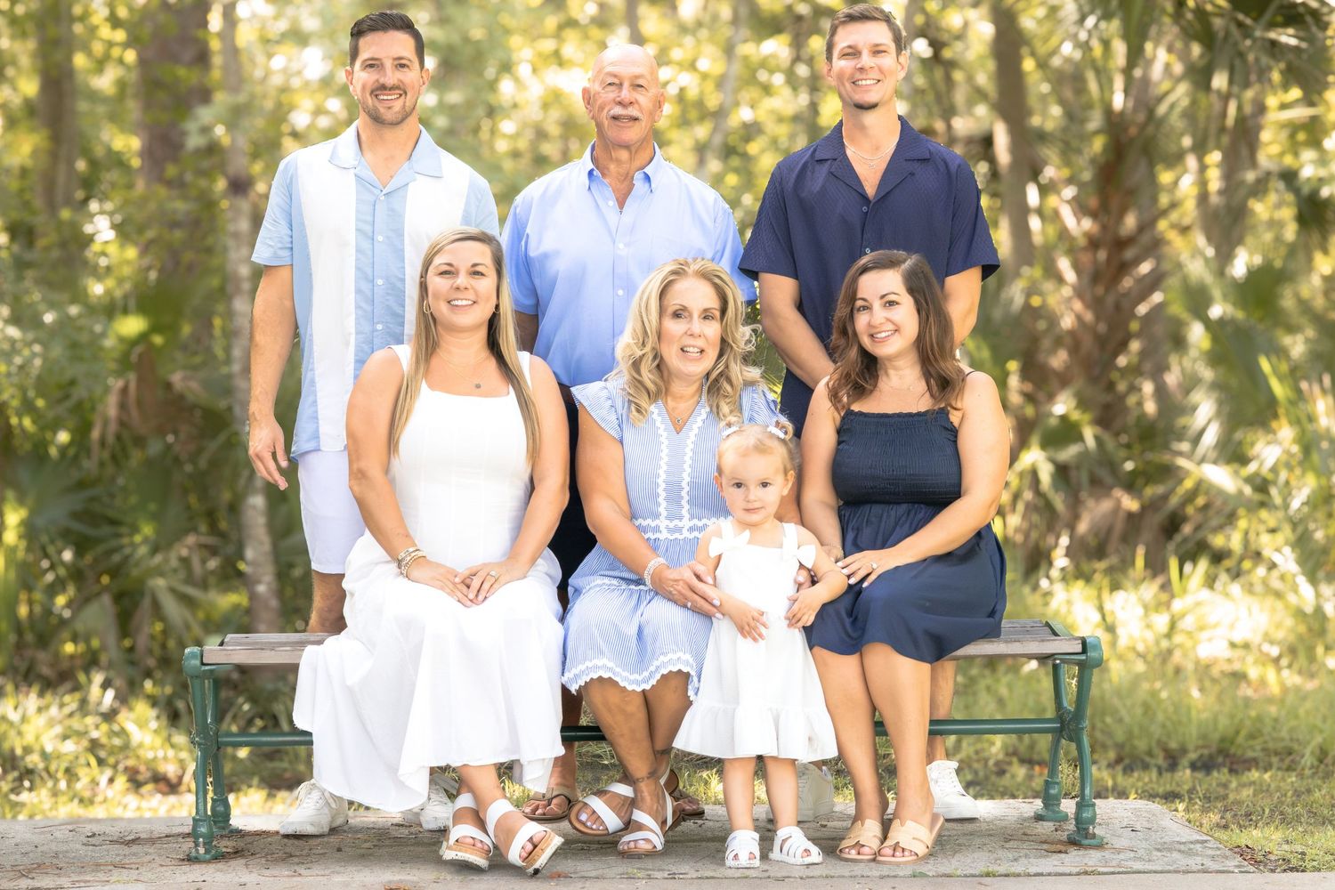 Three generations of a family in coordinated blue and white outfits pose together on a wooden bench in a sunny, tree-filled outdoor setting.