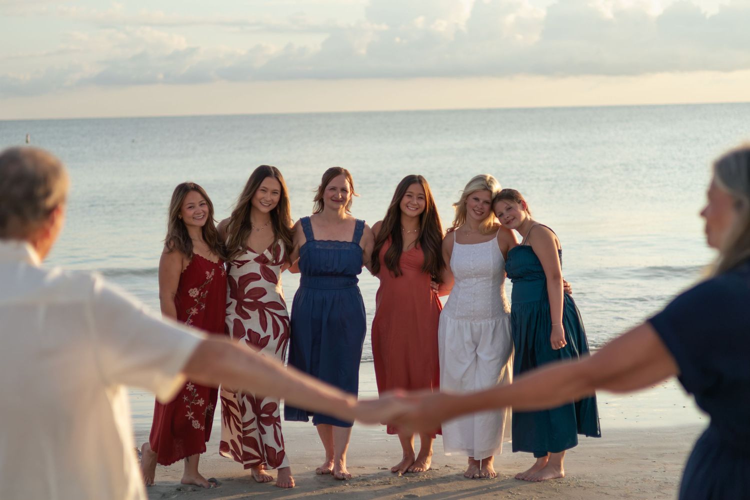 Six women in colorful dresses pose arm in arm on the beach facing the ocean while two family members hold hands in the foreground.