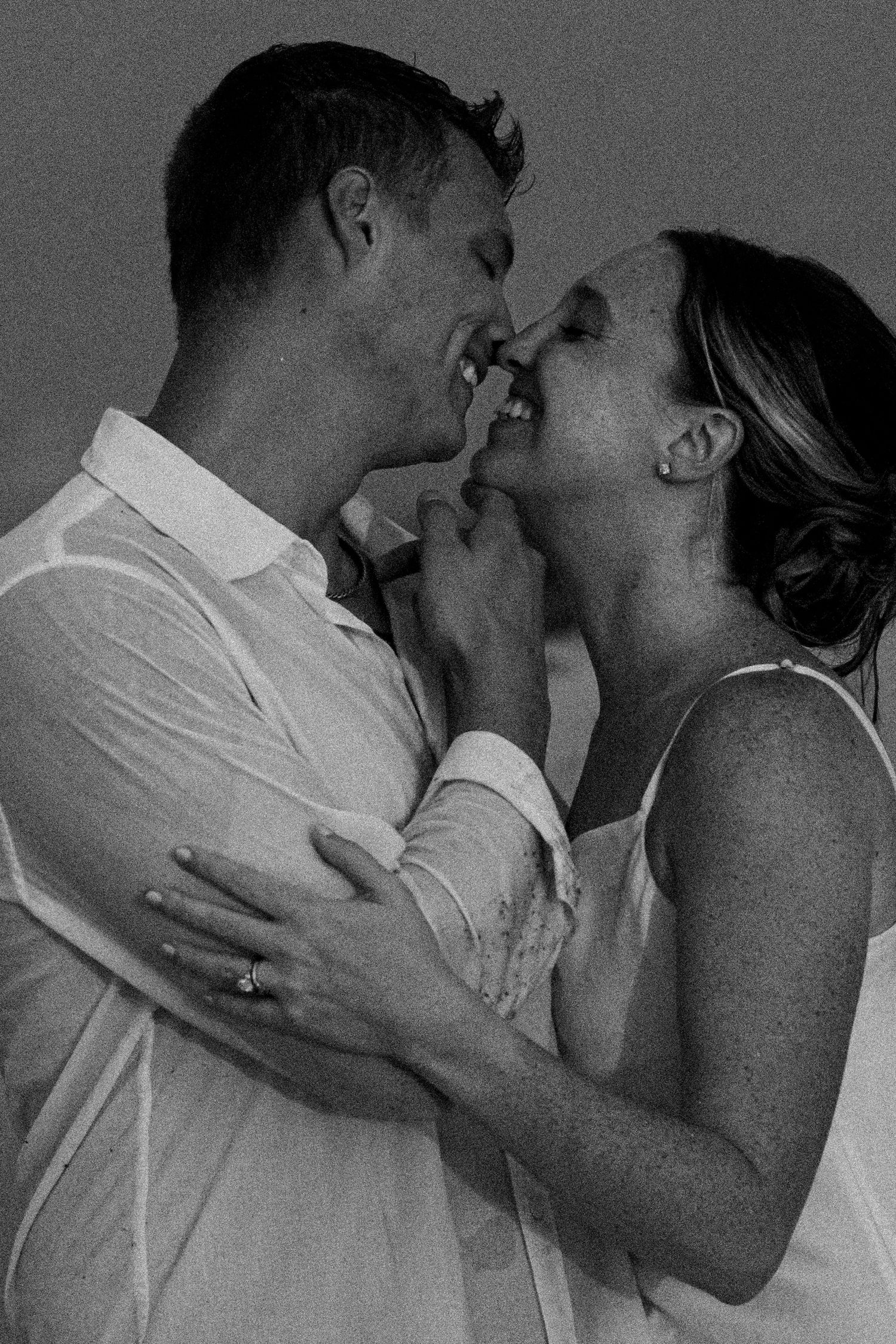 Black and white close-up of a smiling couple embracing nose to nose, the woman's engagement ring visible on her hand.