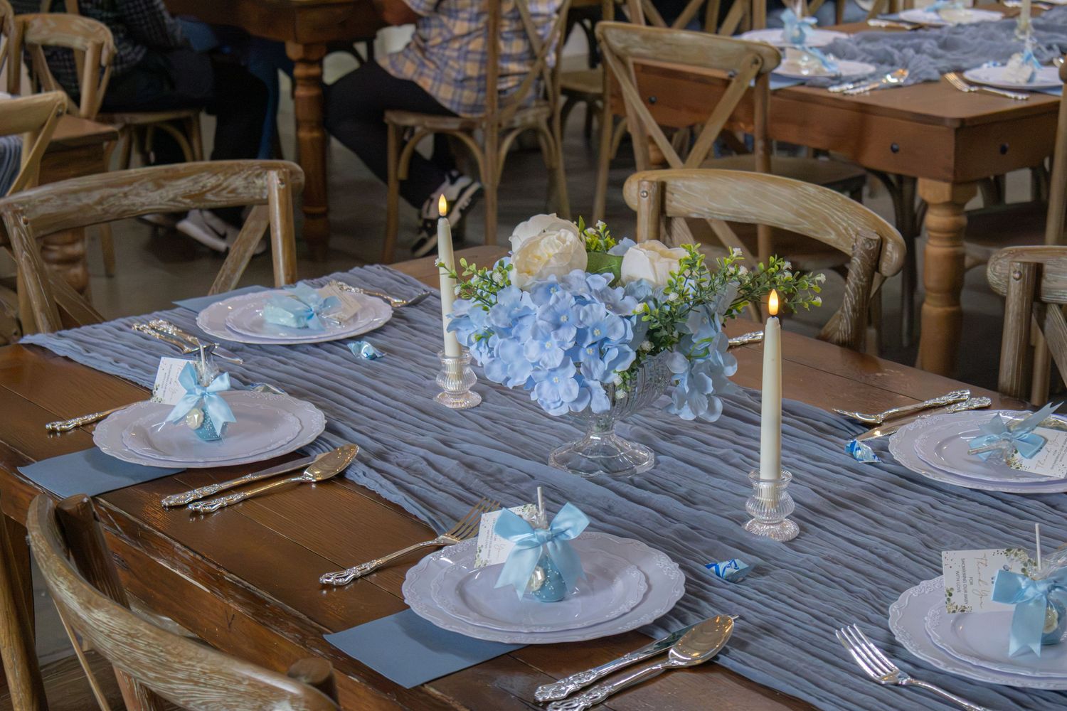 Close-up of a baby shower table with blue hydrangea centerpiece, taper candles and white plates topped with blue bow favors.