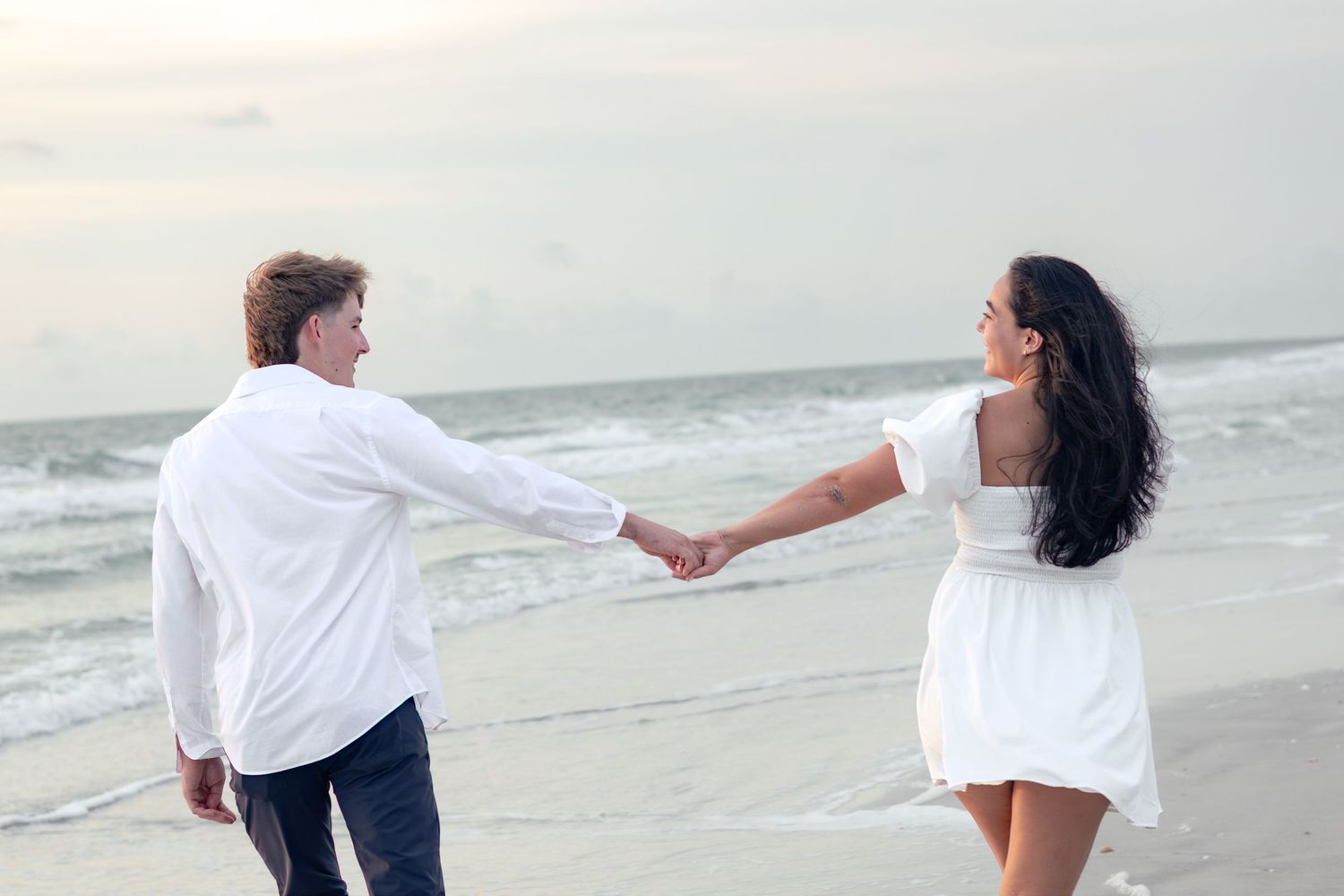 Couple in white outfits holding hands and walking along the shoreline of a calm beach at dusk.