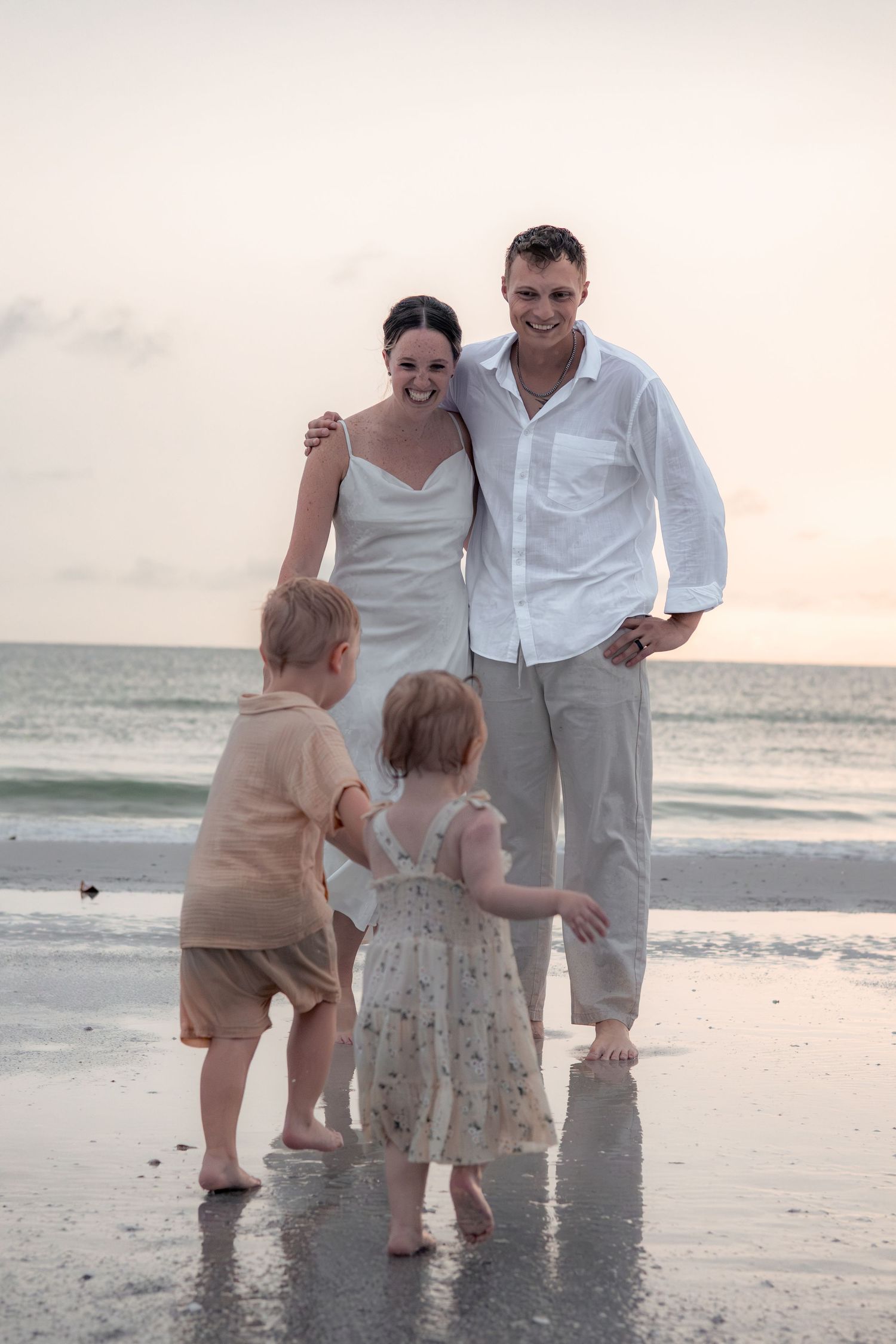 Smiling couple in white outfits laugh together on a beach at sunset while two toddlers play in the shallow water.
