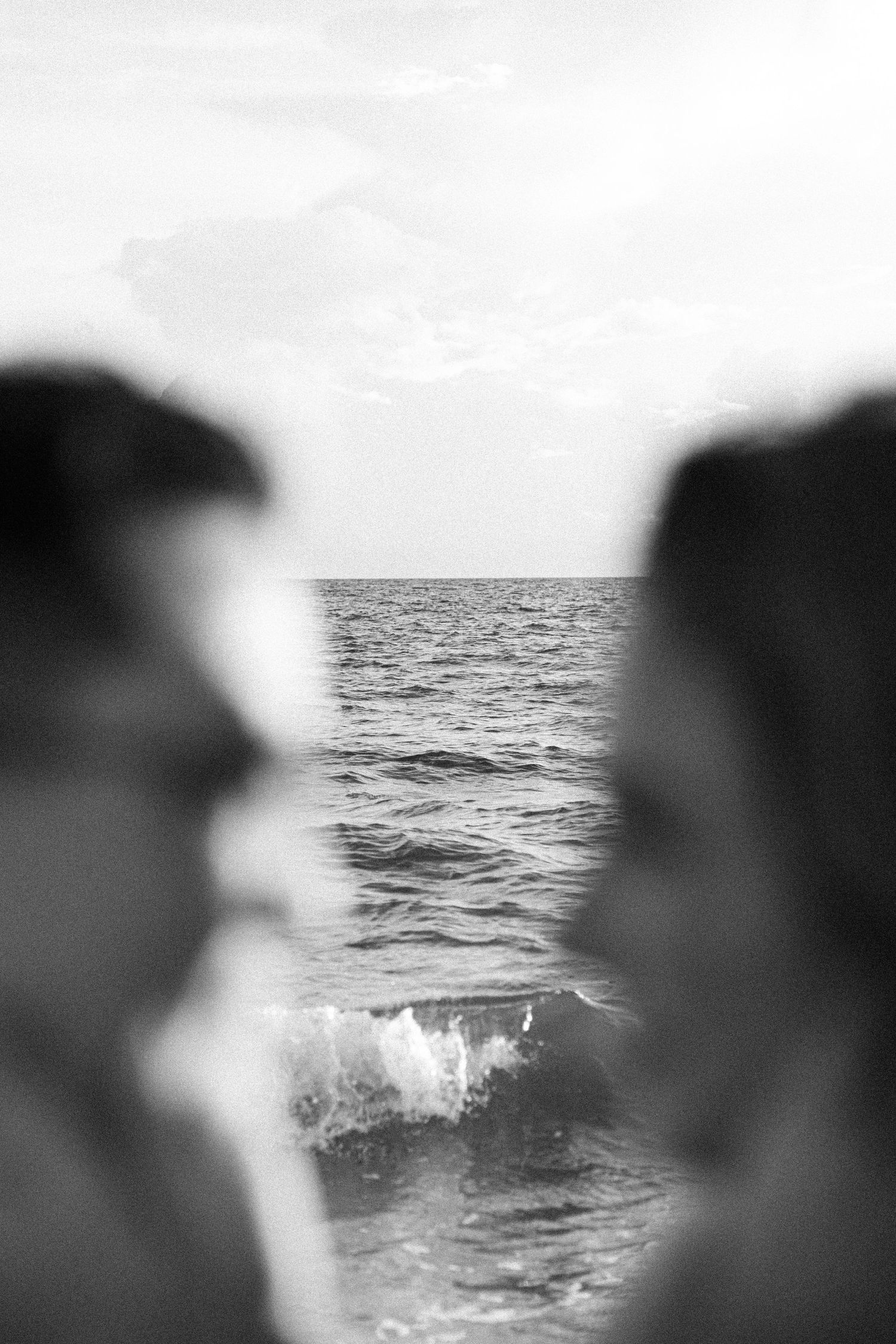 Black and white photo of two blurred faces in profile facing each other, with ocean waves breaking in the sharp background.