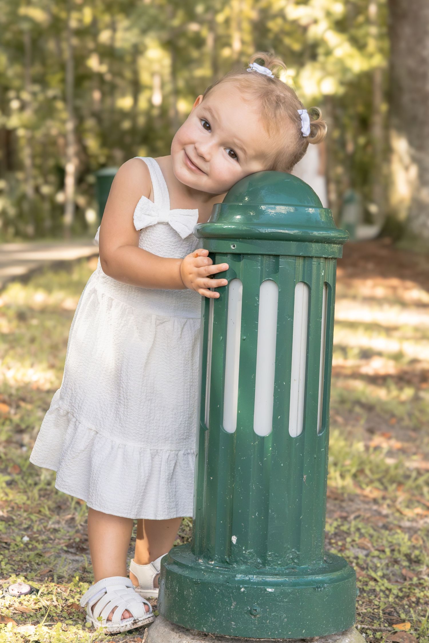 Smiling toddler in a white dress and sandals leans her head on a green park pillar while standing on grass in a sunny, wooded park.